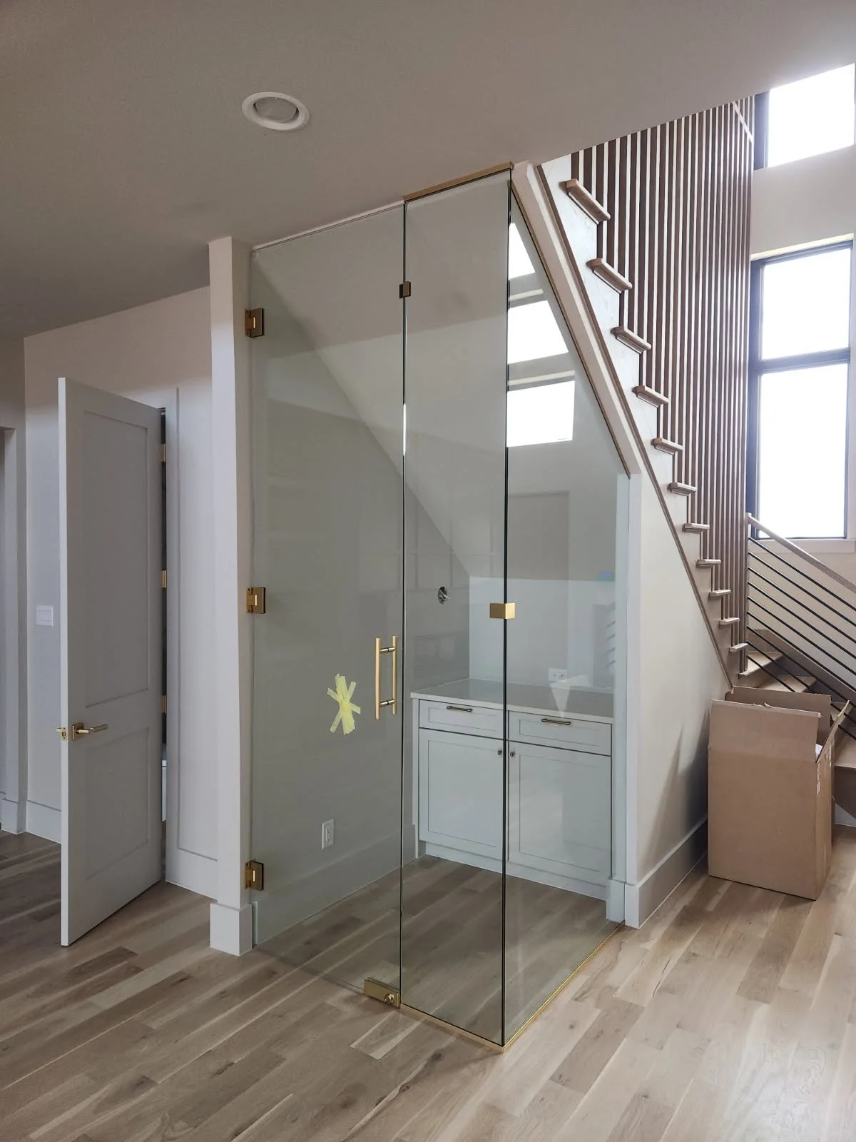 Modern interior space with a glass shower enclosure, white cabinetry, wooden stairs, and large windows in a residential home.