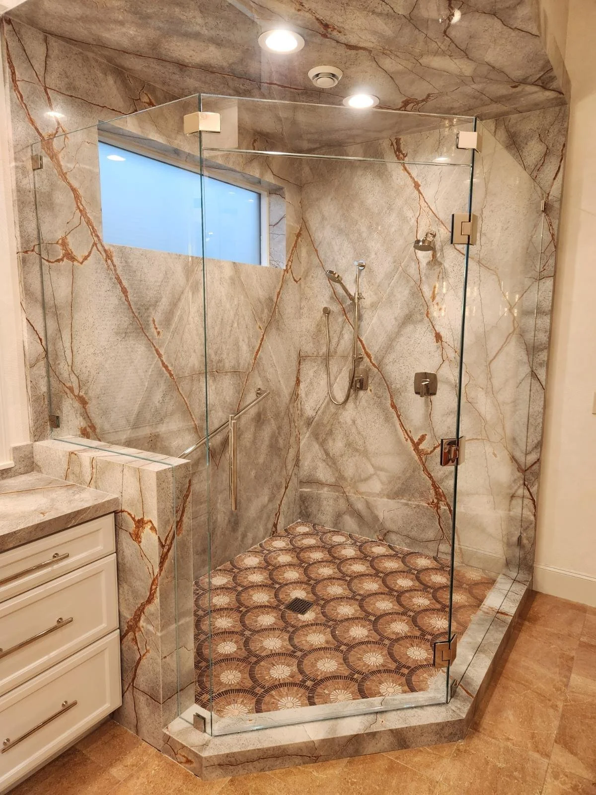 Marble-tiled bathroom shower with glass enclosure, recessed ceiling lights, and patterned floor tiles.