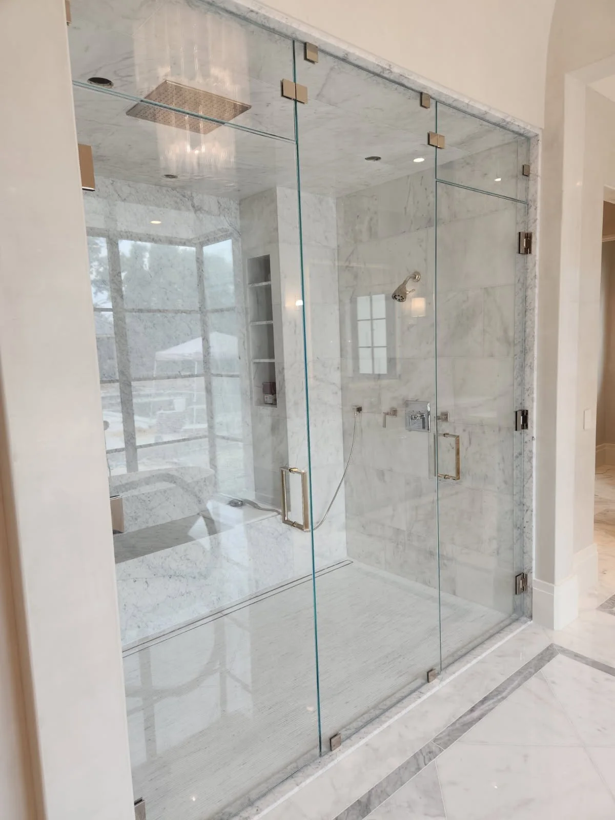 A glass-enclosed shower with marble walls and floor, featuring a ceiling rainfall shower head, a handheld shower head, and a built-in niche for toiletries.