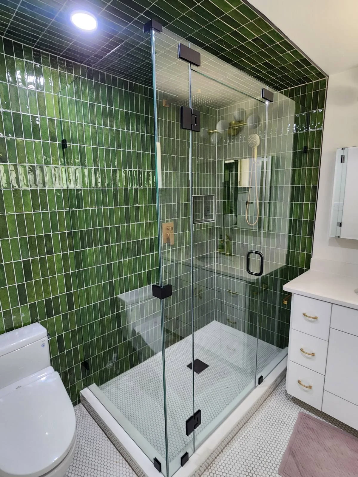 A modern bathroom with a glass-enclosed shower featuring green tile walls, a white toilet, and a white vanity with drawers.