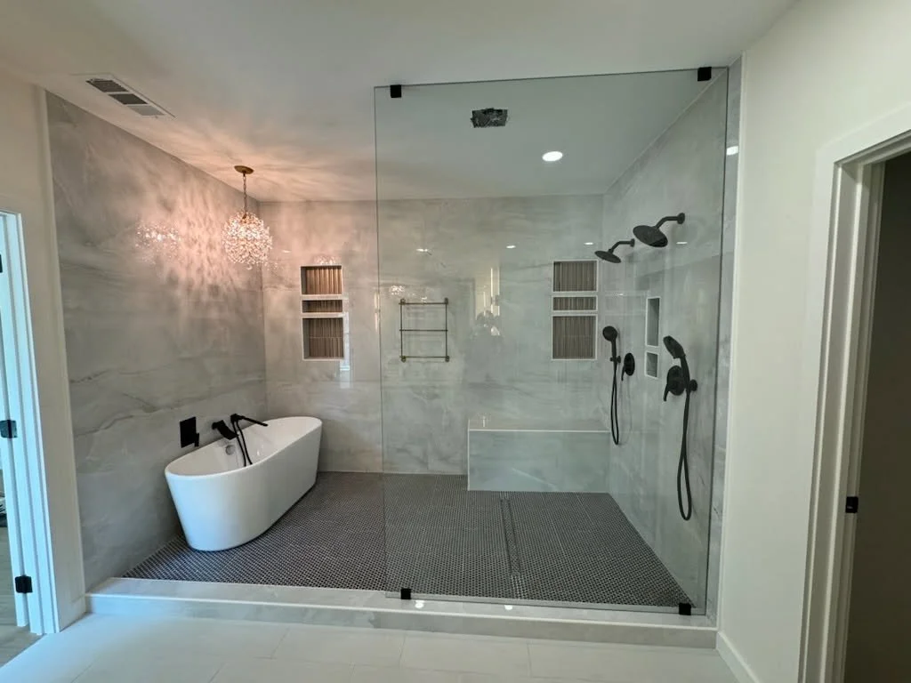 Modern bathroom with a large glass-enclosed shower area containing multiple showerheads and a built-in shelf, and a separate freestanding bathtub with a chandelier overhead.