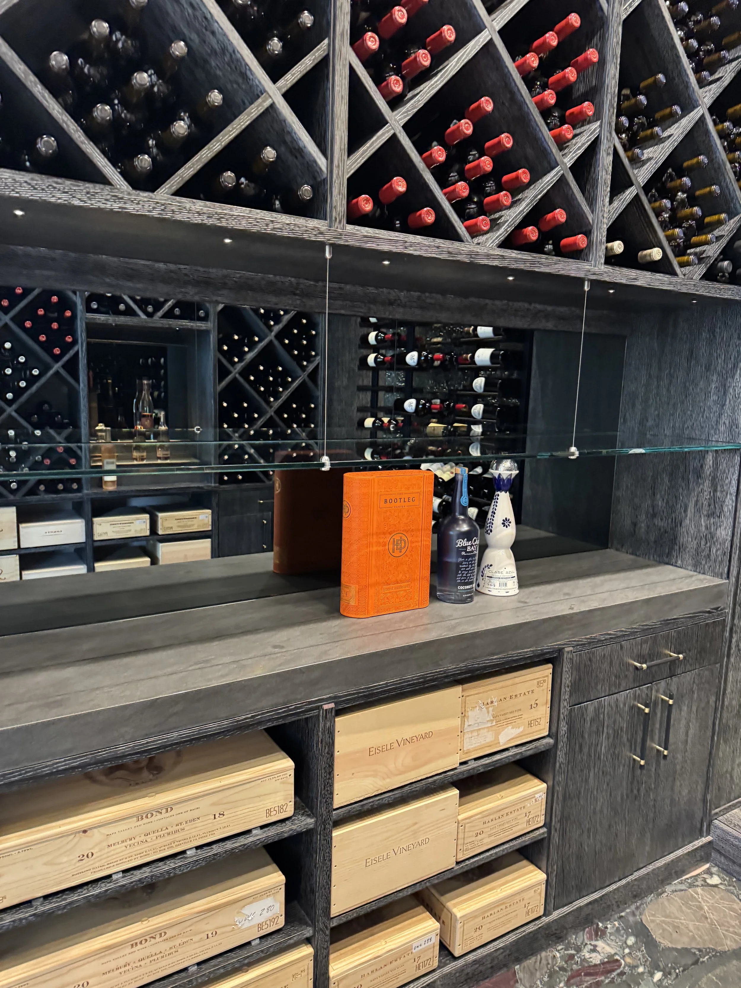 A wine cellar with wooden wine racks filled with bottles, some with red caps, and boxes of wine stored below the racks.