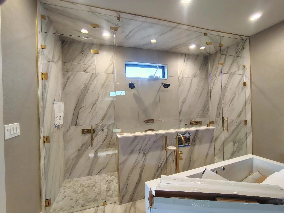A glass-enclosed walk-in shower with marble tile walls and ceiling, two shower heads, small rectangular window, and gold fixtures, in a modern bathroom.