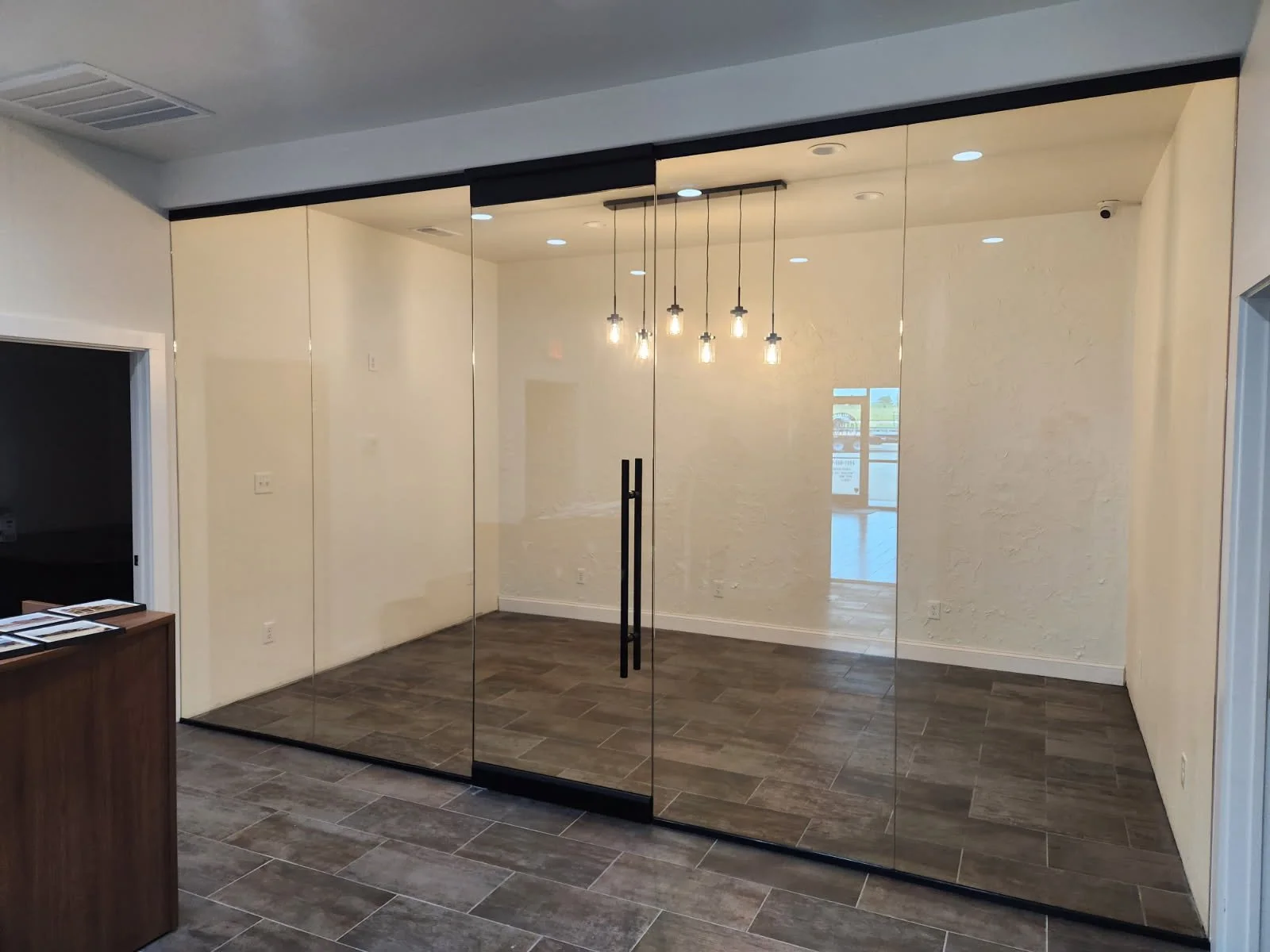 Interior view of a modern office or commercial space with glass double doors, dark wood flooring, and white textured walls, illuminated by pendant lights hanging from the ceiling.
