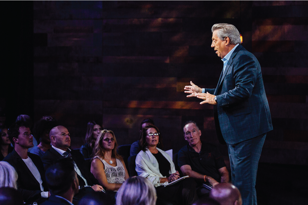 John Maxwell in a blue suit giving a speech or presentation to an audience in a dark room with wooden walls. Leadership Content.