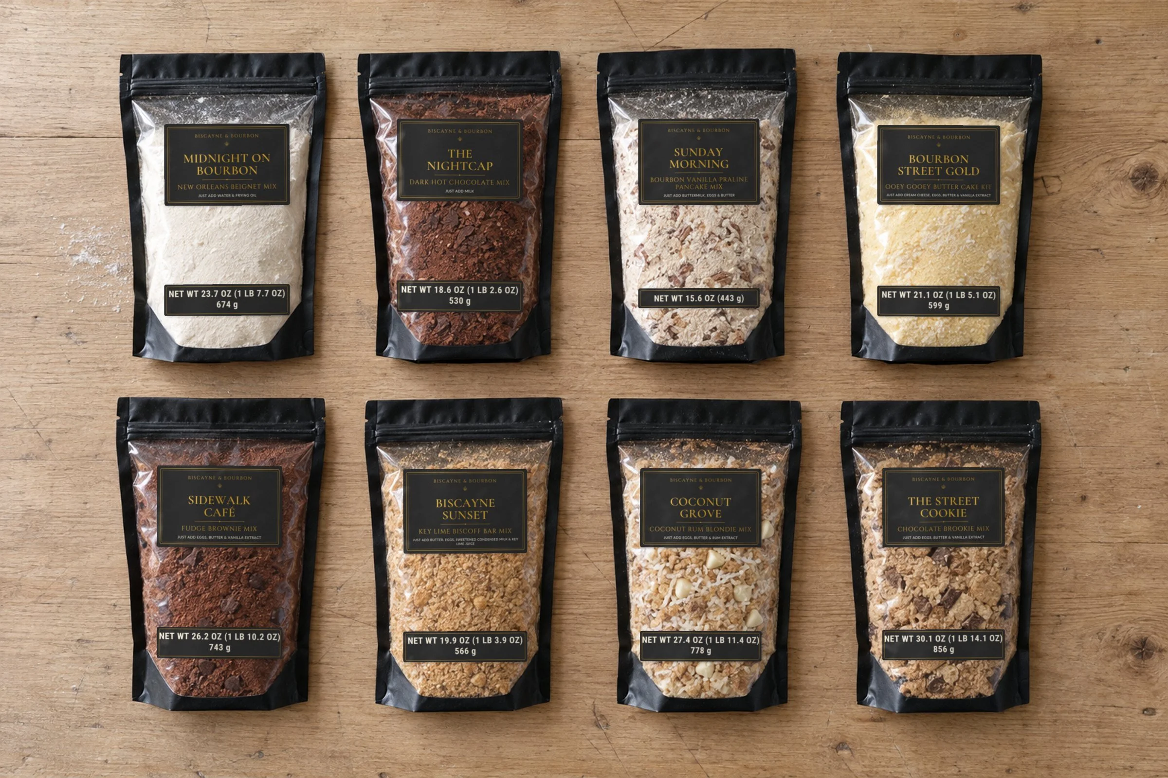 Several bags of baking mixes laid out on a wooden surface, with labels indicating flavors like 'Midnight on Bourbon,' 'The Nightcap,' 'Sunday Morning,' and 'Bourbon Street Gold.'