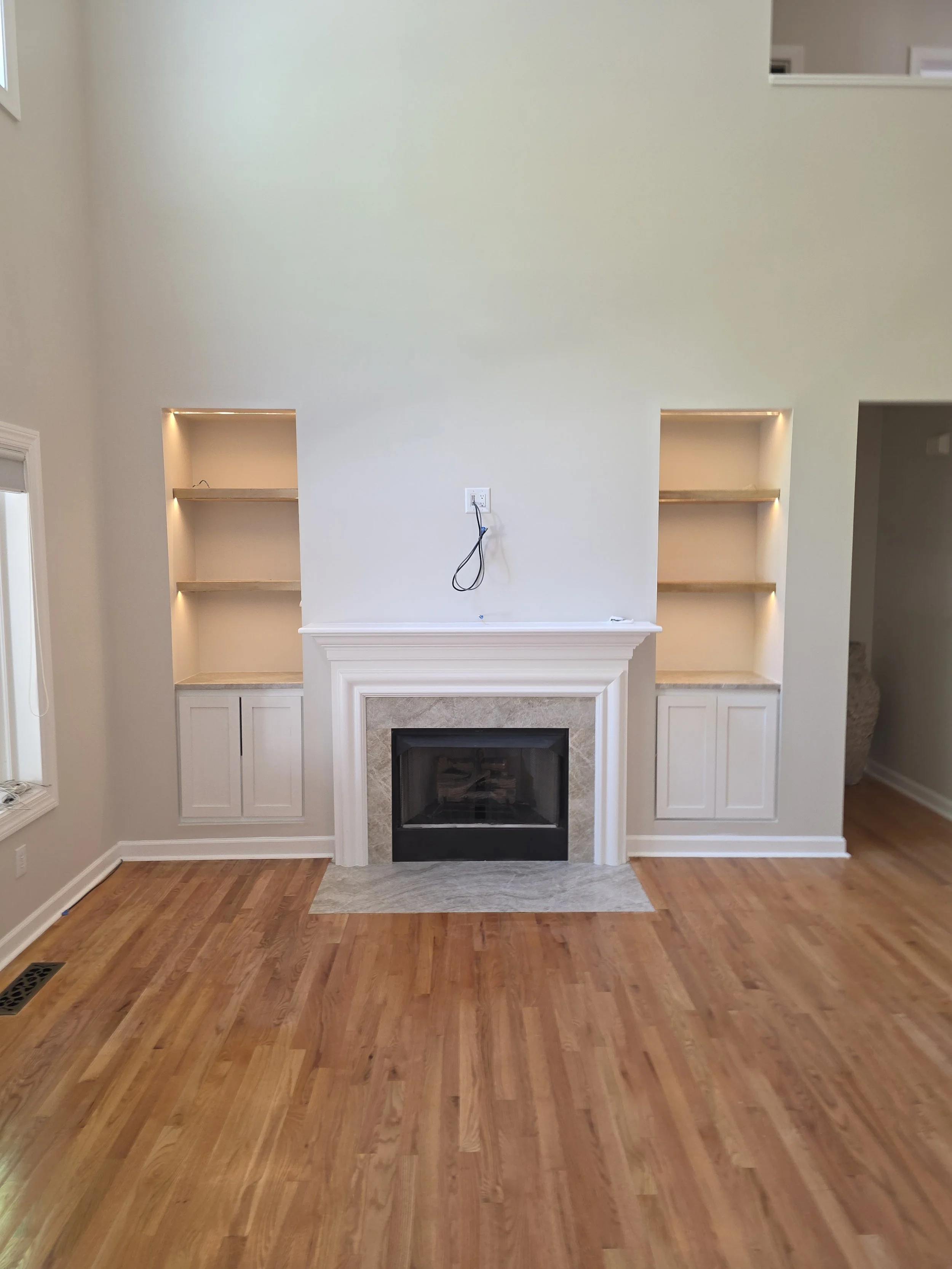 Interior of a house with a fireplace and built-in shelves on either side, with a wall-mounted outlet and cord above the fireplace, and hardwood floors.