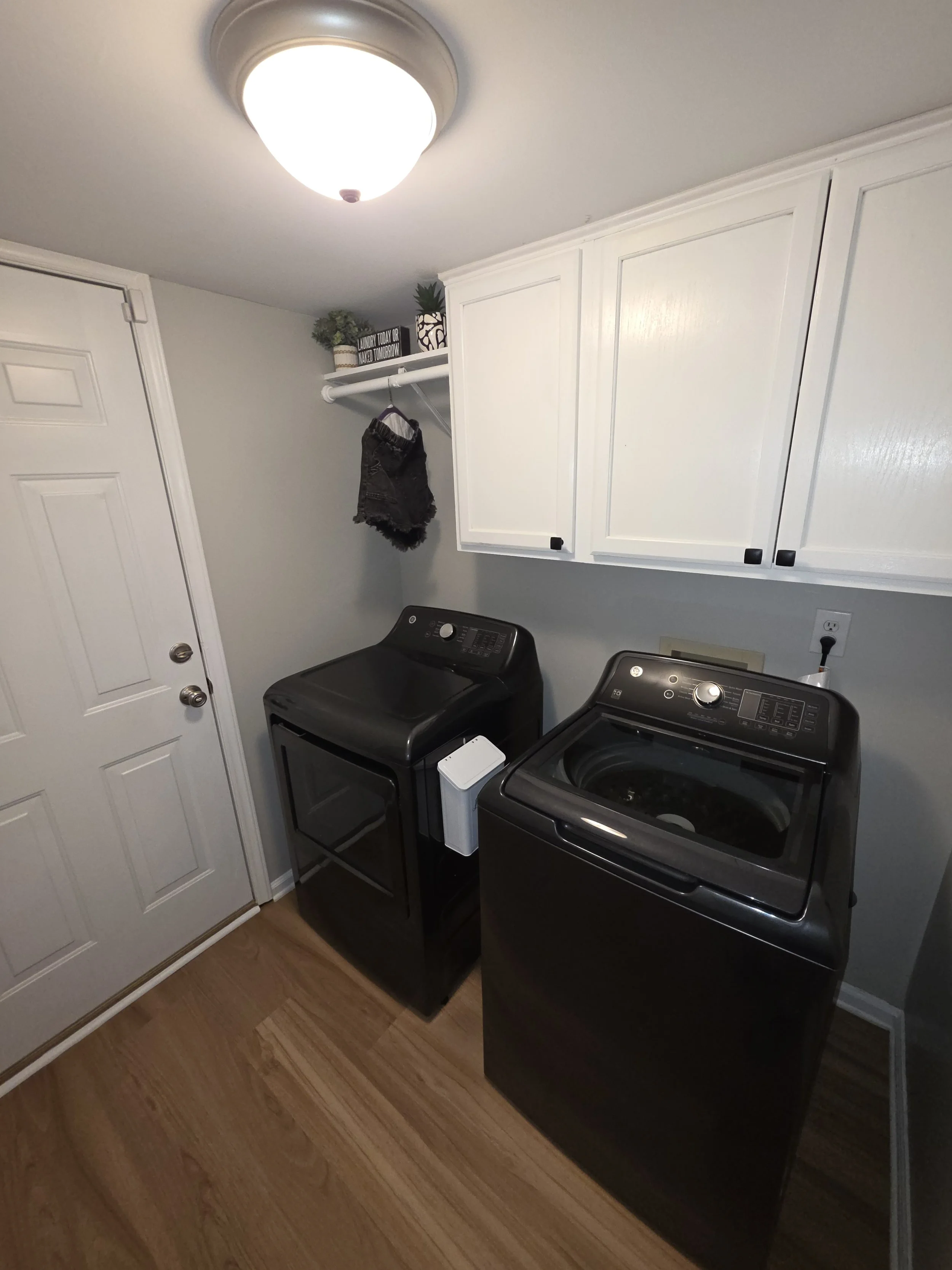 A laundry room with a white cabinet, a front-loading washing machine and dryer, a small side table, and a shelf with decorative plants and a sign that reads 'Laundry today or naked tomorrow.'