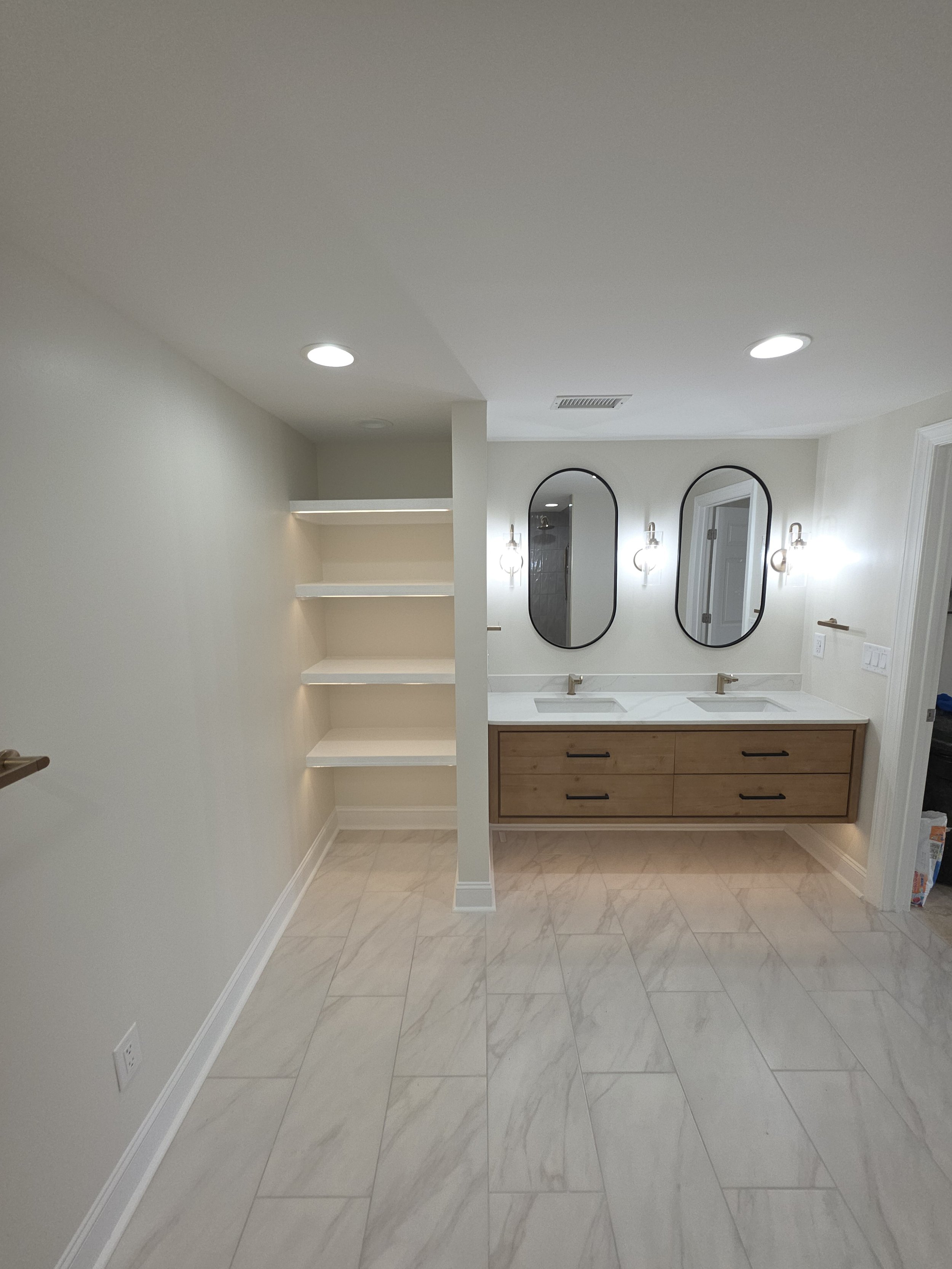 Interior view of a modern bathroom with a double vanity, two oval mirrors, wall-mounted lights, beige walls, and marble flooring.