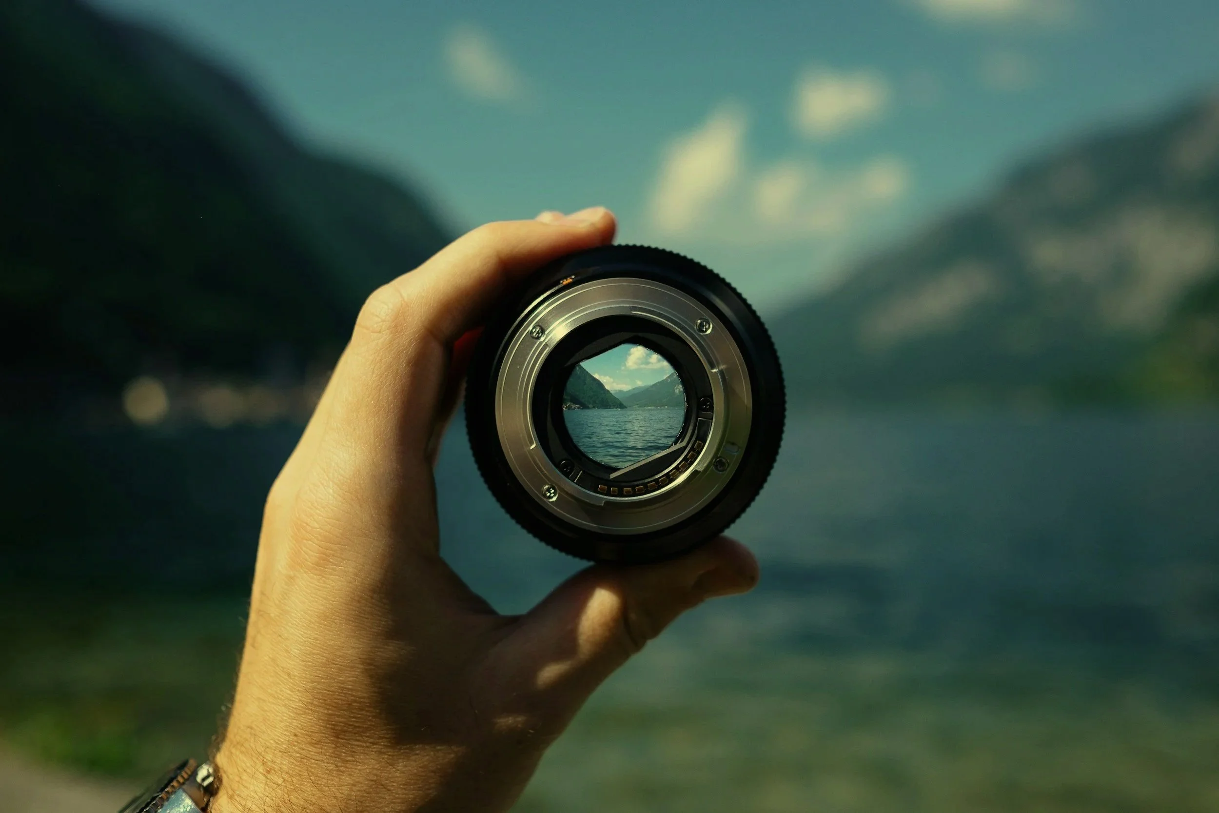 A hand holding a camera lens with a scenic landscape of mountains and water reflected in the glass, under a partly cloudy sky.