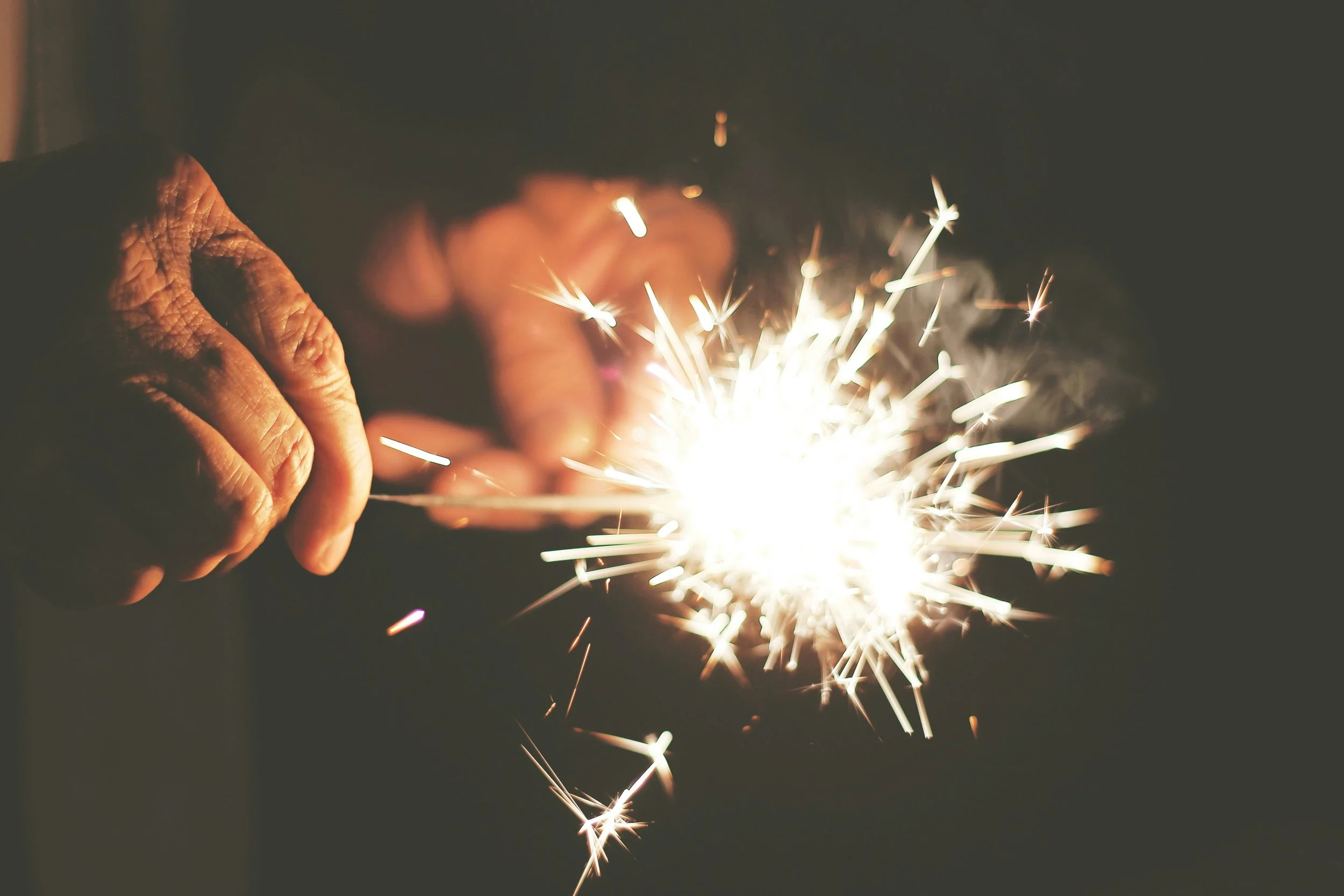 Close-up of hands holding a lit sparkler, with bright sparks flying in a dark background.
