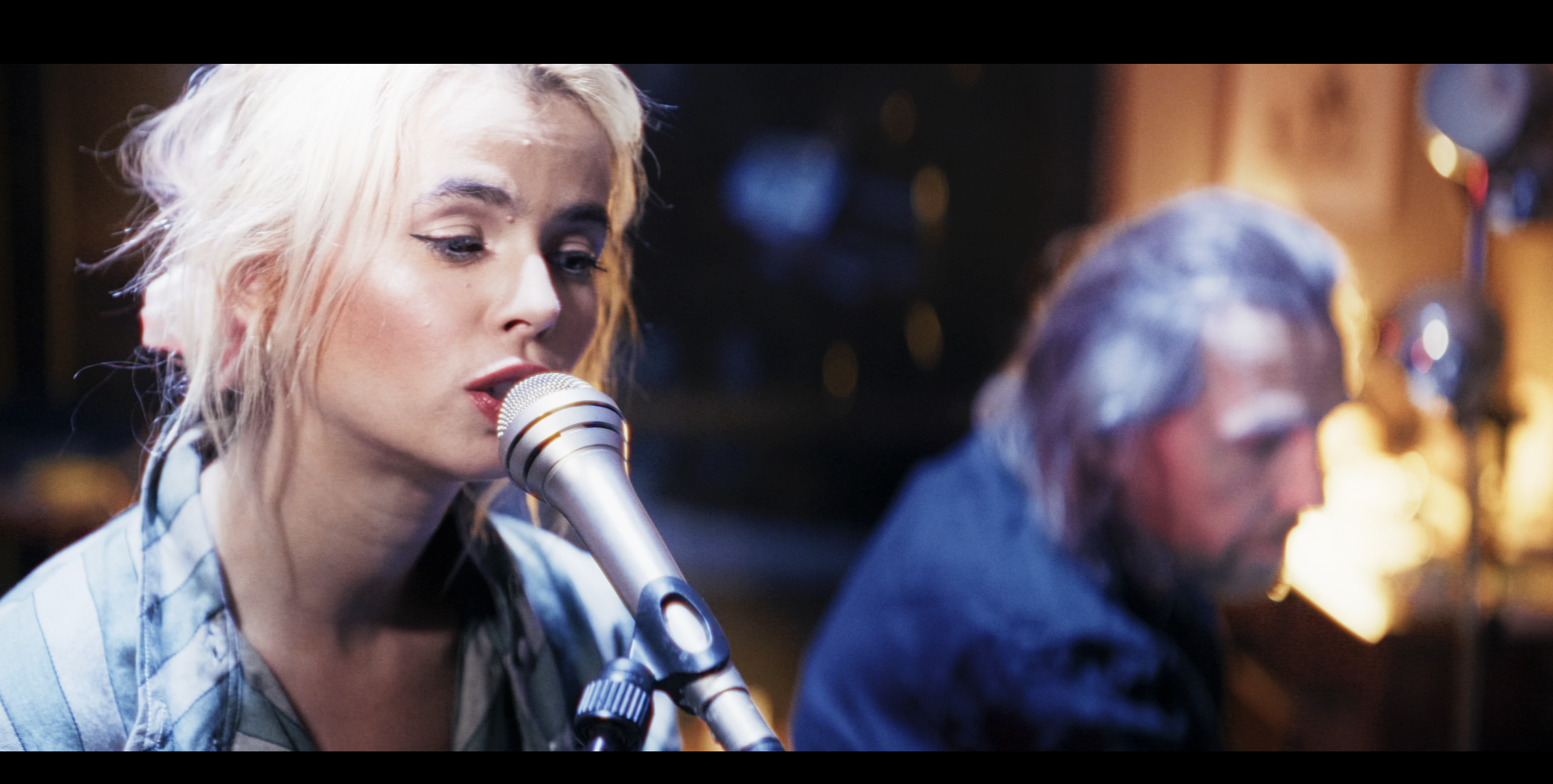 A young woman with platinum blonde hair singing into a microphone in a recording studio, with a man with gray hair in the background.