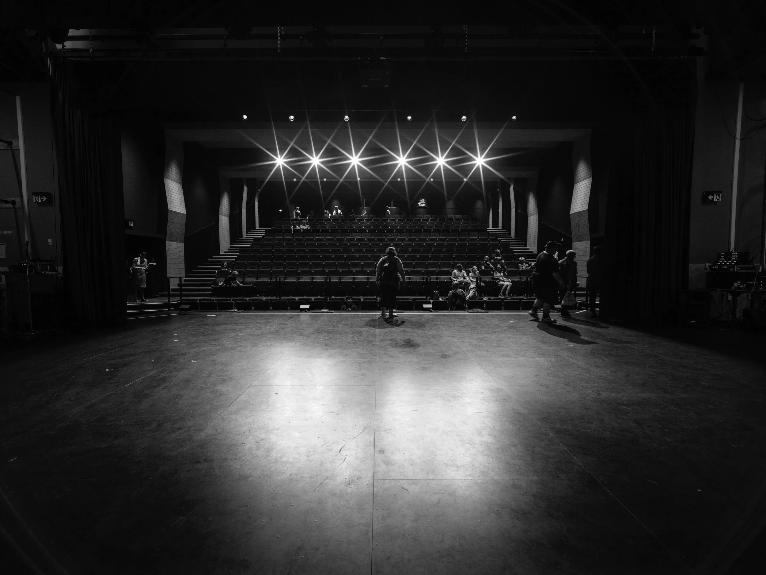 View of an empty theater stage with a dark background and bright stage lights shining down, with some people sitting and standing in the audience area.