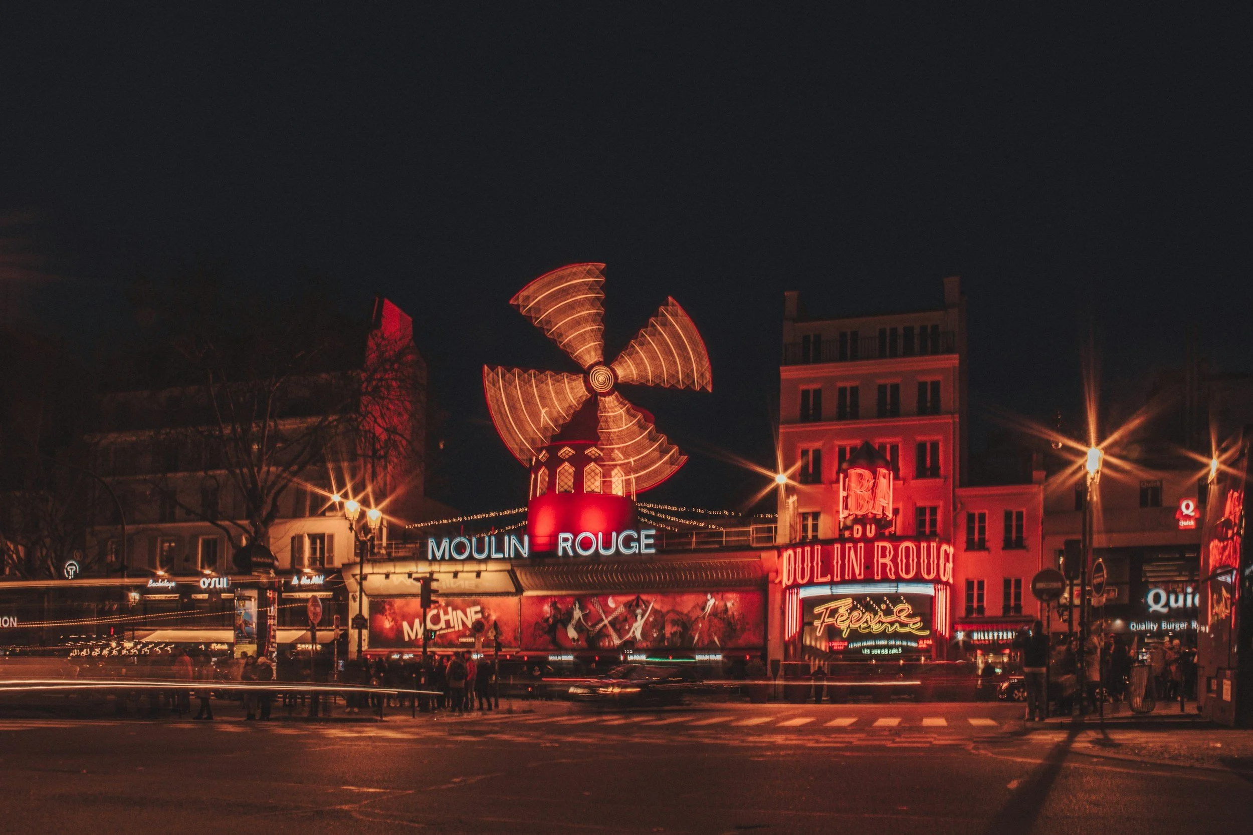 Night view of the Moulin Rouge in Paris with vibrant red neon signage and rotating windmill, illuminated buildings, and street scene.