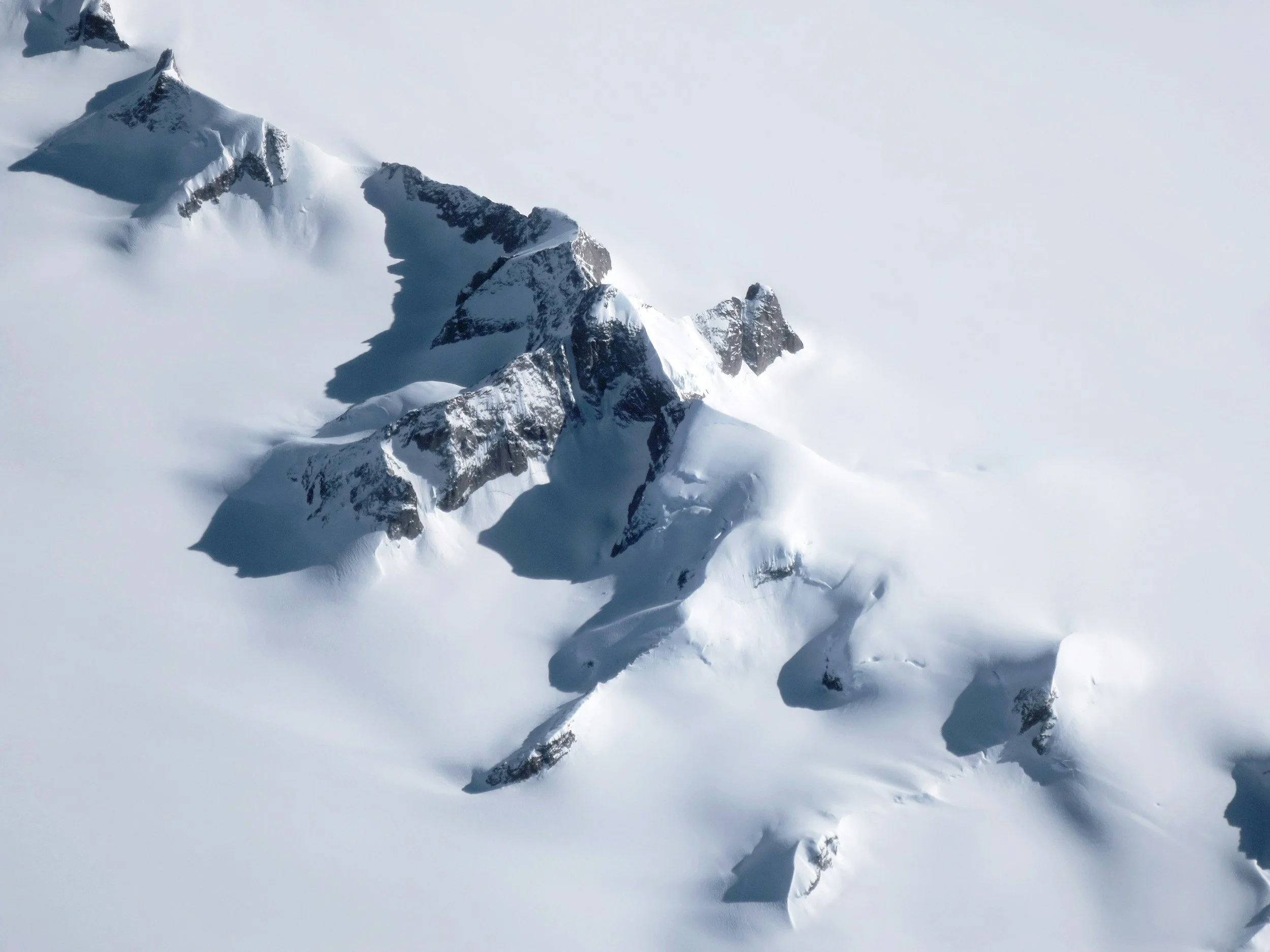 Snow-covered mountain peaks with jagged, dark rocks protruding through the snow, under a pale, cloudy sky.