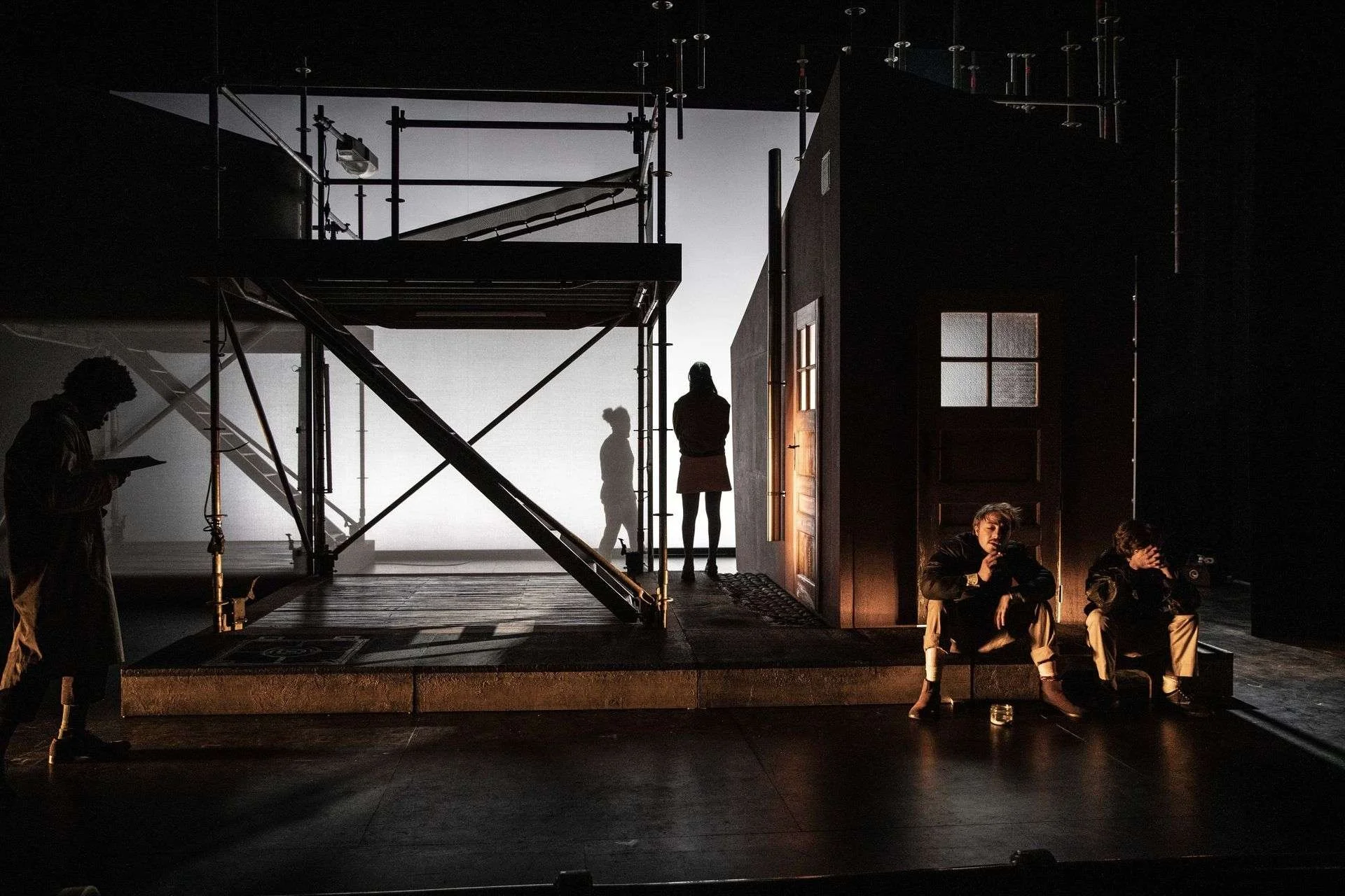 Behind the scenes of a theatrical or film production, with silhouettes of crew members and actors on stage, scaffolding, and stage equipment, illuminated by dim lighting.