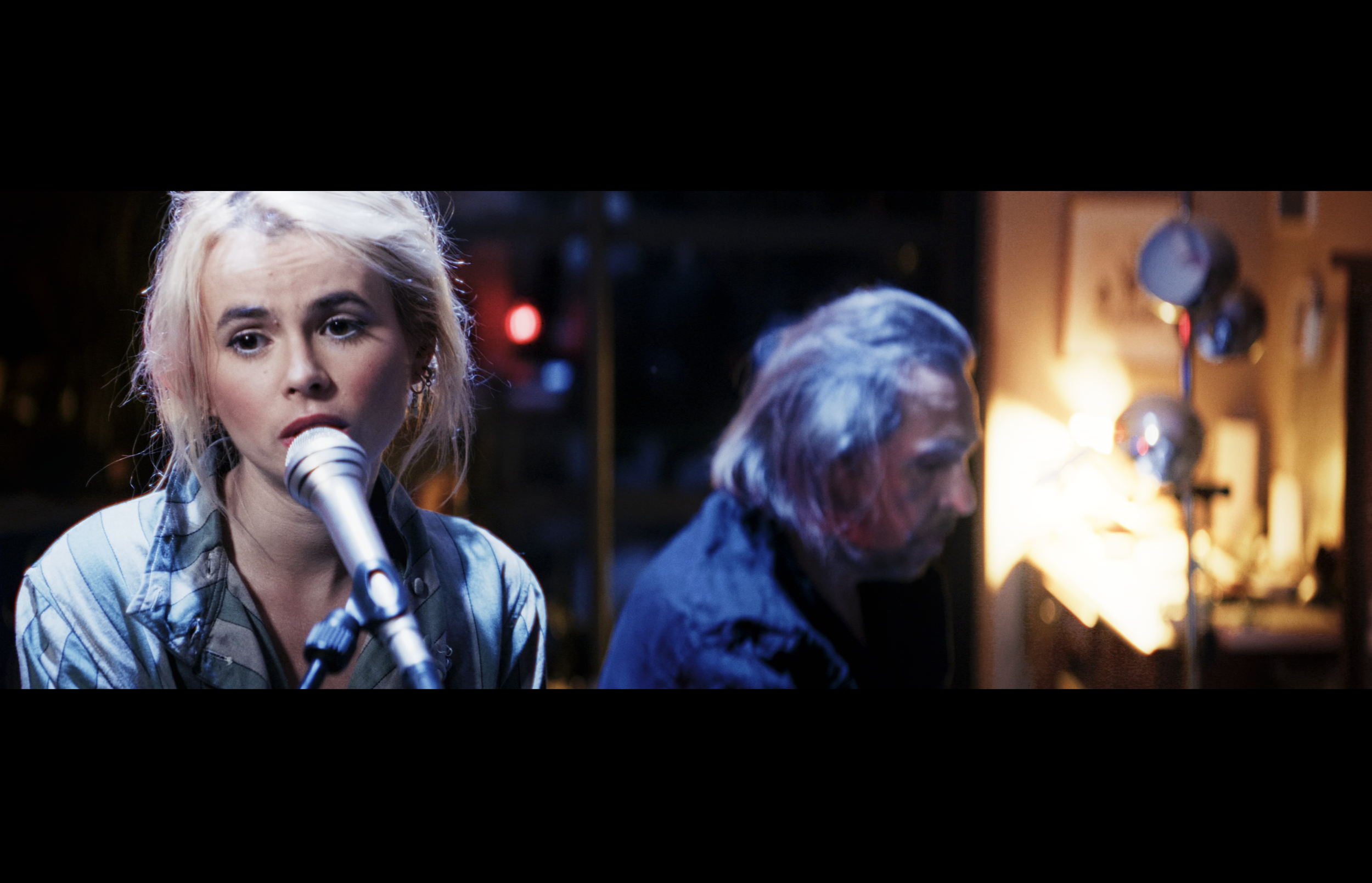 A young woman with blonde hair singing into a microphone, wearing a striped shirt, in a dimly lit room with another person playing a keyboard nearby.