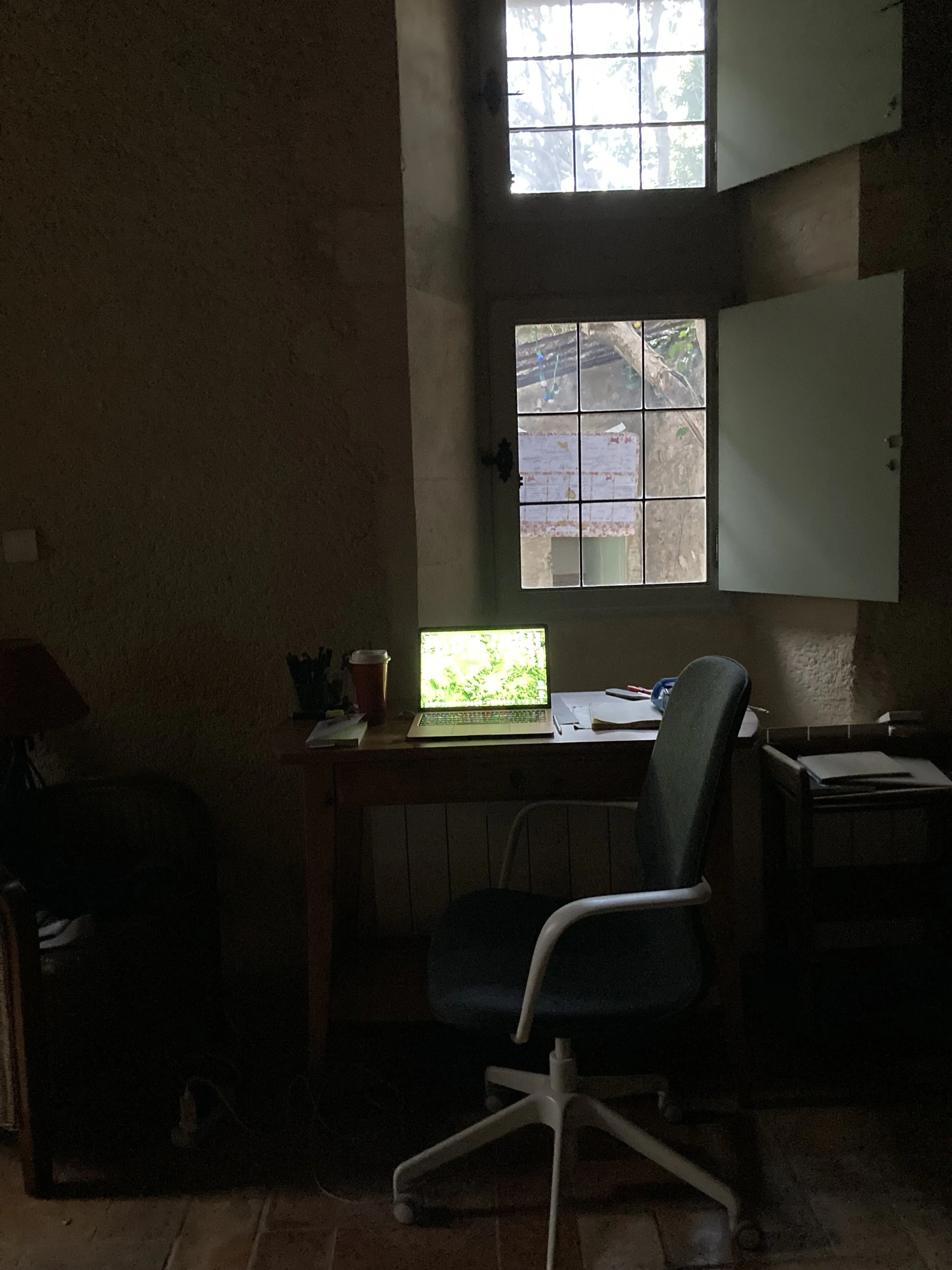 A dimly lit room with two windows, one open and one closed, showing trees outside. A wooden desk with a laptop, papers, and pens is in front of the windows. An office chair with a white and black frame is in front of the desk. To the left, another chair and some items are visible, with a tiled floor.