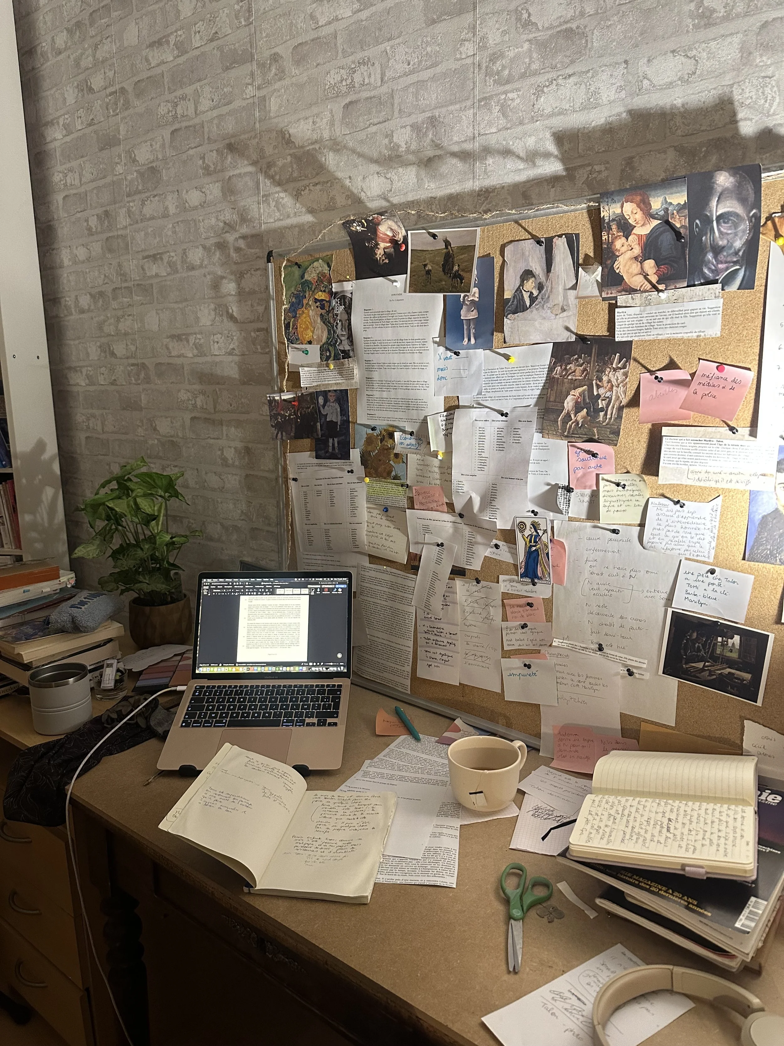 A cluttered study desk with an open laptop, notebooks, pens, a cup, and scattered papers. Behind the desk is a cork bulletin board filled with various photographs, notes, and documents pinned up, with a plant on the left side.