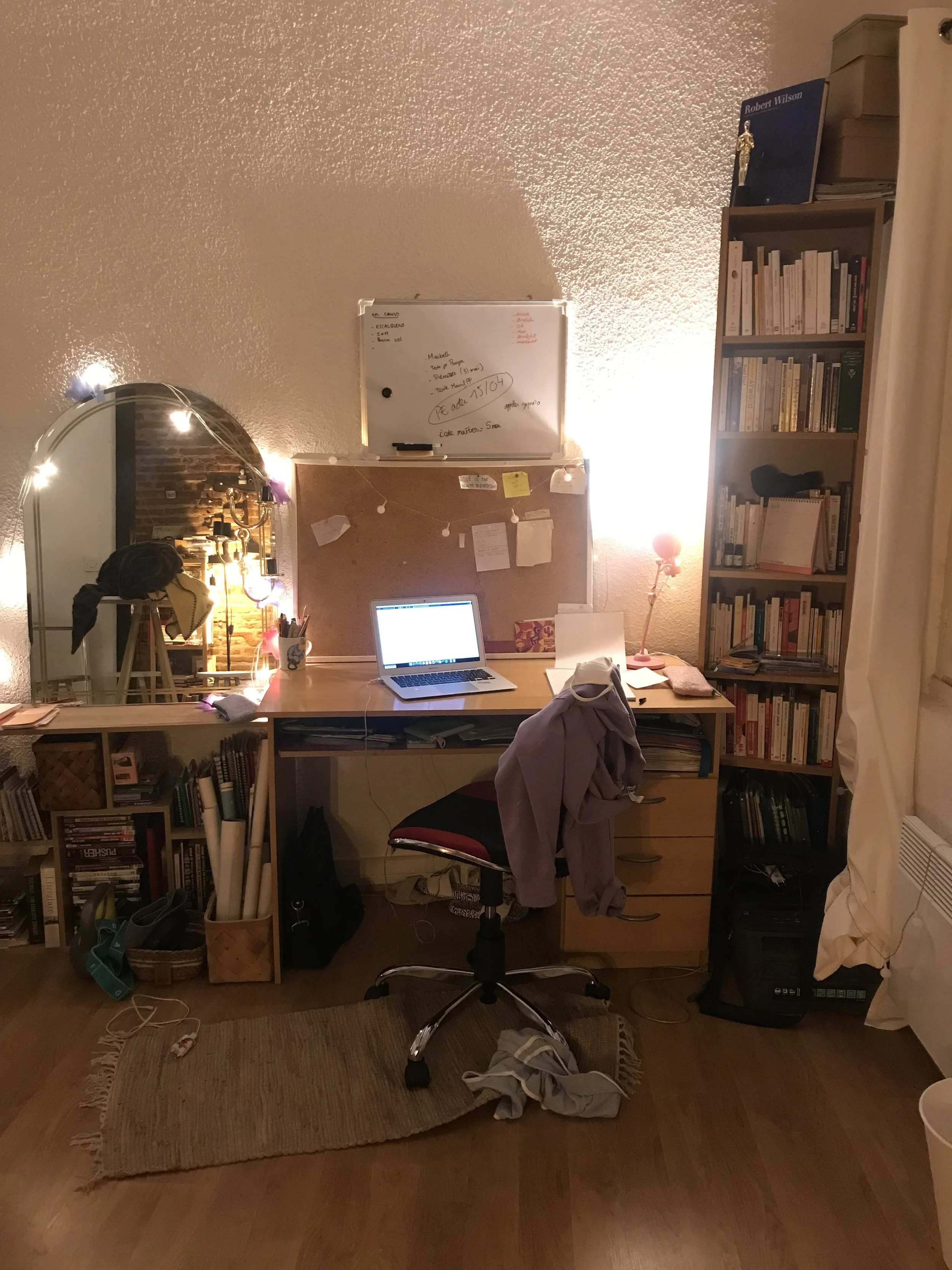 A cluttered home office desk with a laptop, a pink lamp, a whiteboard, and a bookshelf filled with books. The room has string lights, a mirror, and various items including a chair with a coat, papers, and shoes.