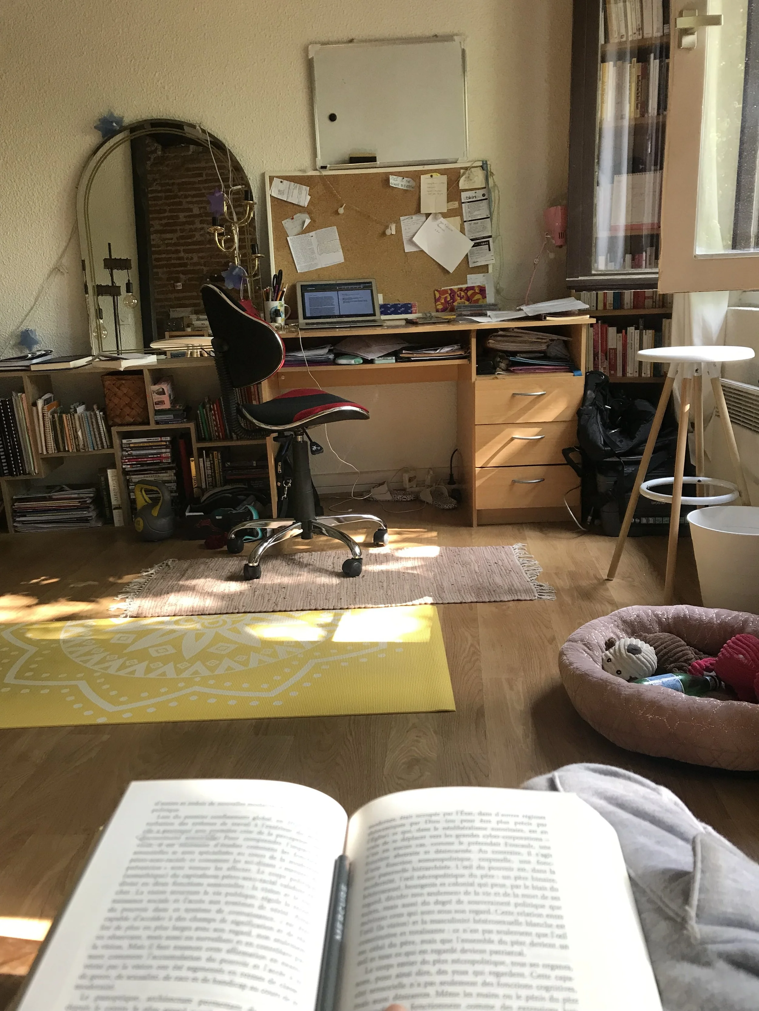 Photo of a cozy home workspace with a person reading a book. The room features a cluttered desk with a laptop, papers, and a bookshelf filled with books. A window lets in natural light, and a small round table with a stool is nearby. The foreground shows the person's lap and the open book.