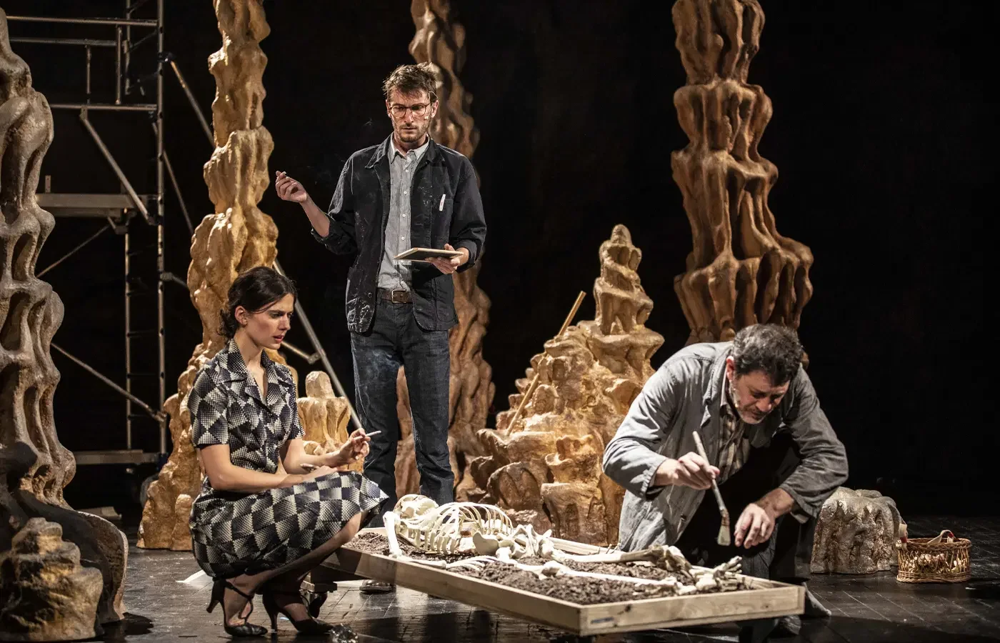 Three actors on stage with dinosaur skeletons and fossils, one woman squatting, one man standing, another man kneeling, engaged in a theatrical performance.