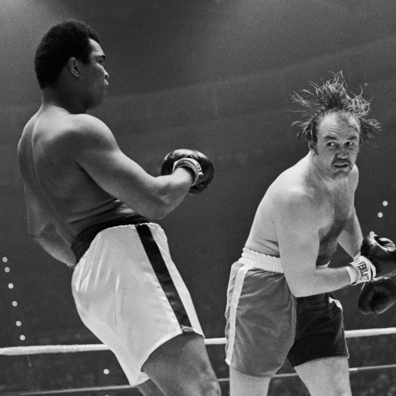 Black and white photo of two male boxers in a boxing match, one delivering a punch to the other. The boxer on the left has a darker uniform and is wearing boxing gloves, while the boxer on the right has disheveled hair and appears to be reacting to the punch.