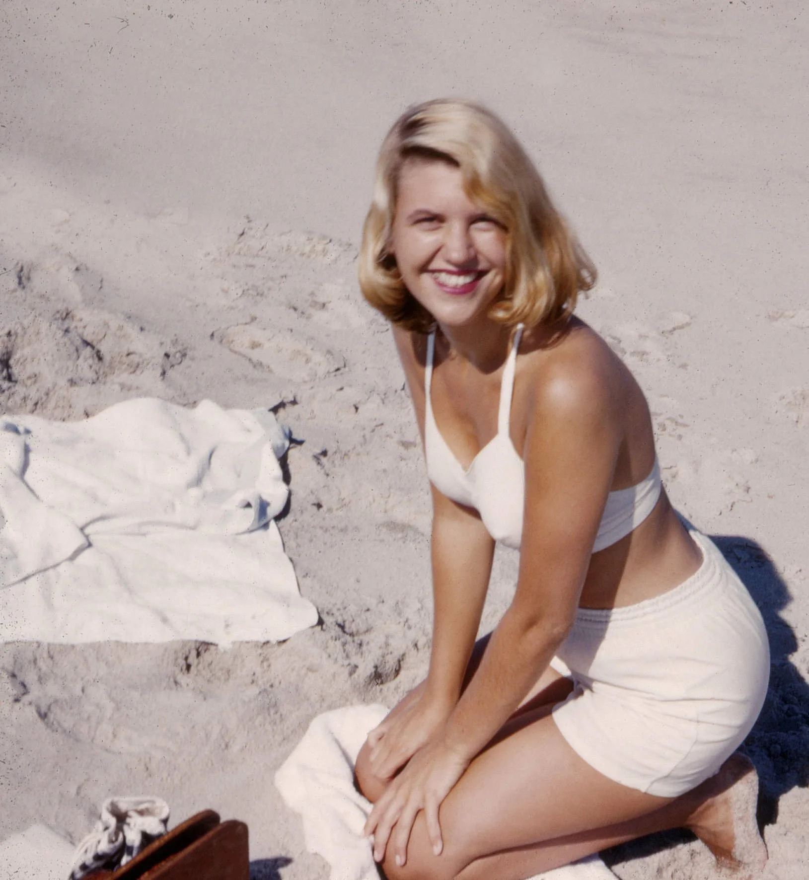 Woman with shoulder-length blond hair smiling and crouching on sandy beach, dressed in a white sleeveless top and beige shorts, with a white towel nearby and some shoes in the lower left corner.
