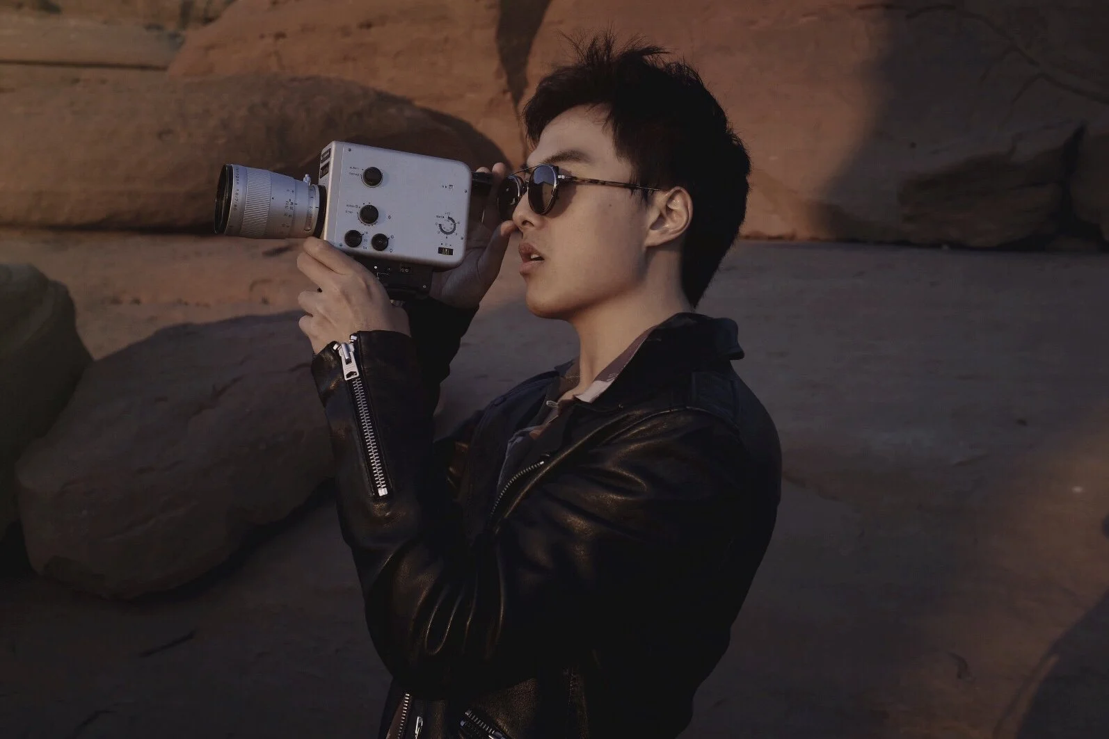 A young man with dark hair wearing sunglasses and a black leather jacket is holding a vintage film camera and looking through its viewfinder against a rocky outdoor landscape at dusk or dawn.