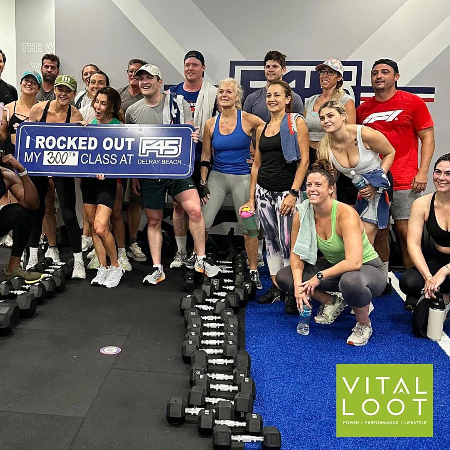 Group of people in workout gear stand together in a gym, holding a large sign that says 'I ROCKED OUT MY 300TH CLASS AT F45 DELRAY BEACH.' There are dumbbells on the floor, and a sign with 'VITAL LOOt' and the words 'Power, Performance, Lifestyle' in the corner.
