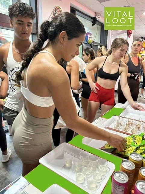 A woman pouring a green drink into small clear cups at a booth with a Vital Loot sign, with people in workout clothes around her.