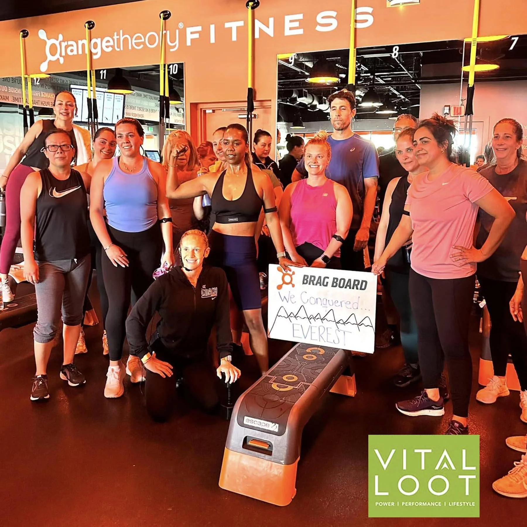 A group of women and a man in workout attire at OrangeTheory Fitness, posing for a photo. They are standing in front of fitness equipment and a sign that says 'BRAG BOARD.' The gym has an orange and black color scheme. There is a green sign in the bottom right corner that says 'VITAL LOOT' with the words 'POWER | PERFORMANCE | LIFESTYLE' underneath.
