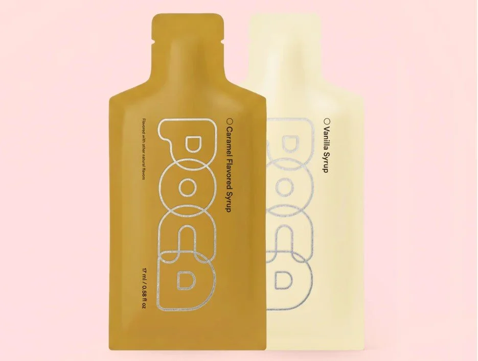 Two bottles of Pod Organic Flavored Syrup, one caramel and one vanilla, on a pastel pink background.