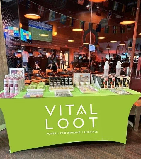 Table with Vital Loot health supplements at an indoor event or gym, with people and exercise equipment visible through the glass background.