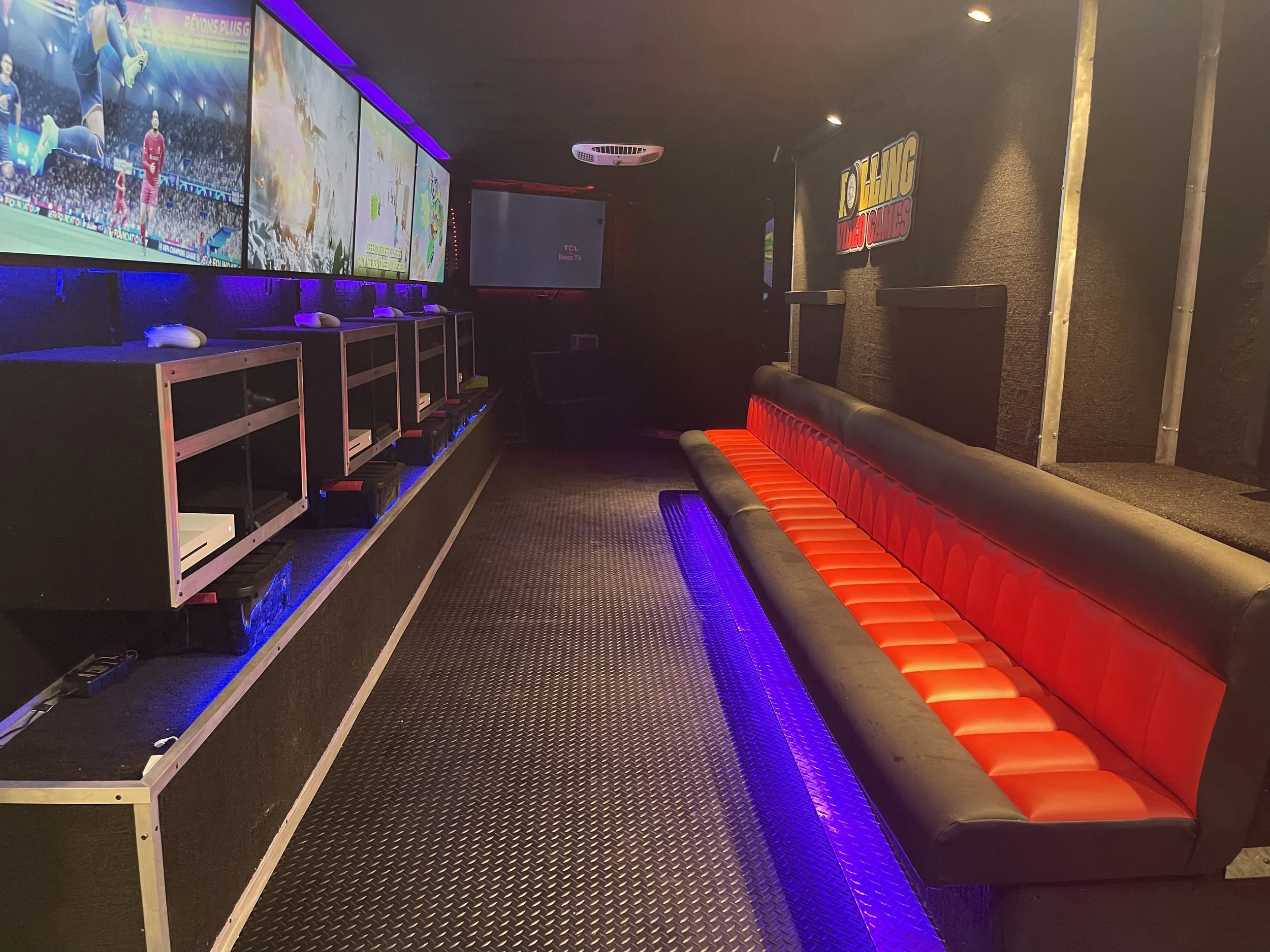 Interior of a mobile gaming trailer with multiple video game screens, gaming consoles, seating and colorful LED lighting.