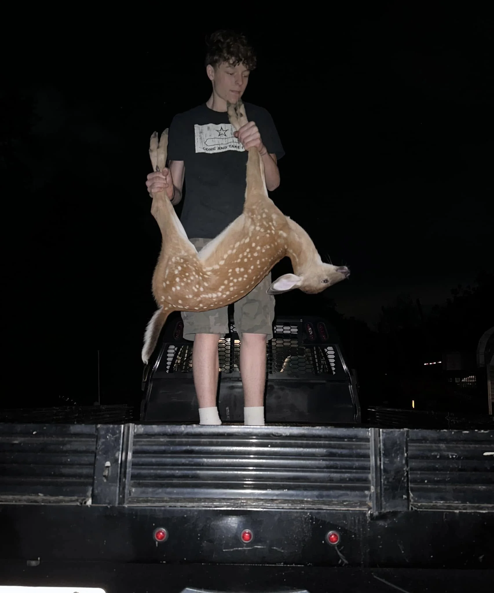 A young man standing on the tailgate of a pickup truck at night, holding a fawn by its hind legs and chest while the deer is upside down with its head near the ground.