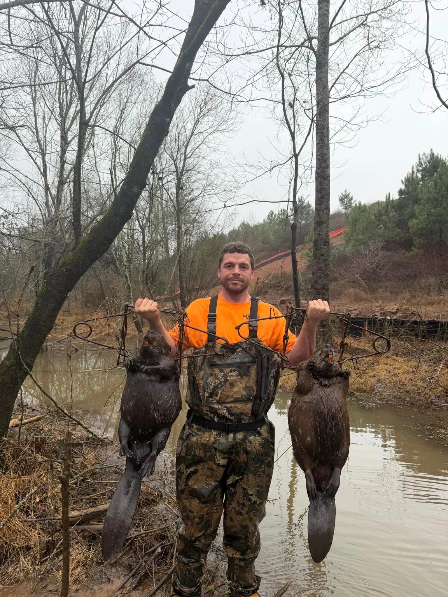 A man standing in a creek holding a fishing net with two fish caught in it. The man is wearing camouflage pants and an orange shirt. There are leafless trees and a small hill in the background.