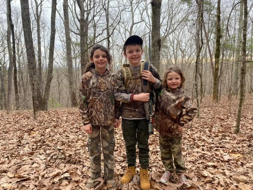 Three children in camouflage clothes standing in a leaf-covered forest, smiling at the camera, with trees and overcast sky in the background.