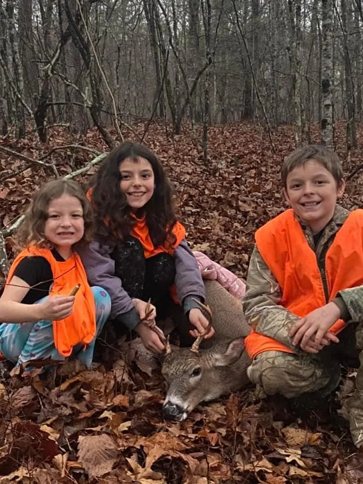 Three children in orange vests posing with a deer they hunted in a forest with fallen leaves and trees in the background.