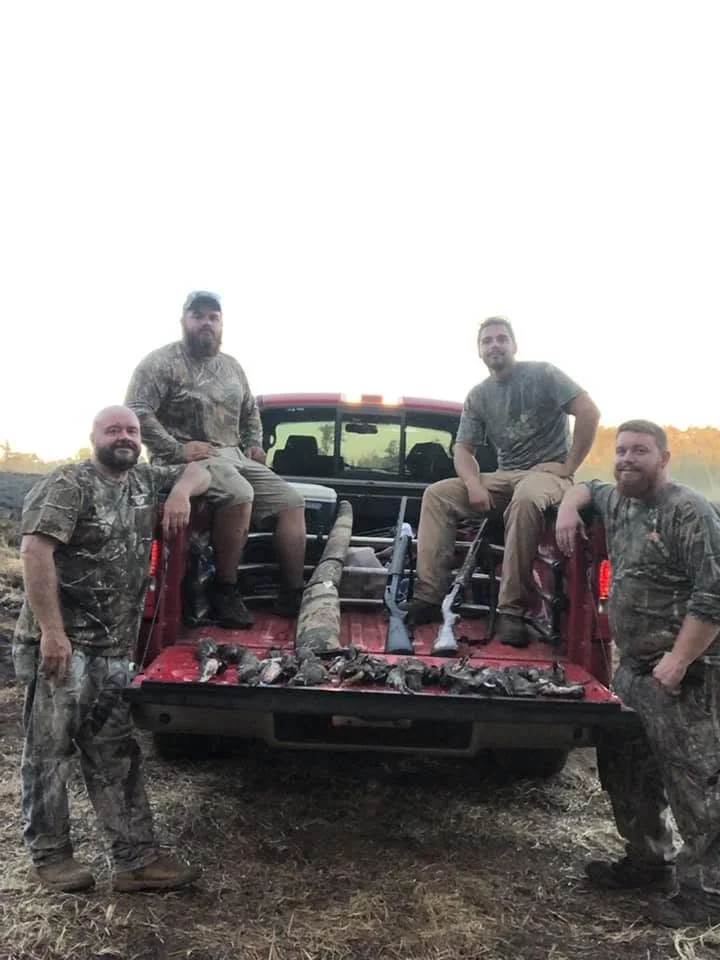 Group of five men in camouflage clothing posing with a truck filled with dead animals, likely during a hunting trip, outdoors during evening or early morning.