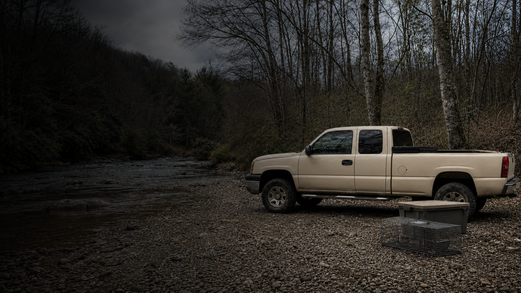A beige pickup truck parked on a rocky riverbank in a wooded area during overcast weather. There are containers and a cage on the ground near the truck, with leafless trees and a flowing river in the background.