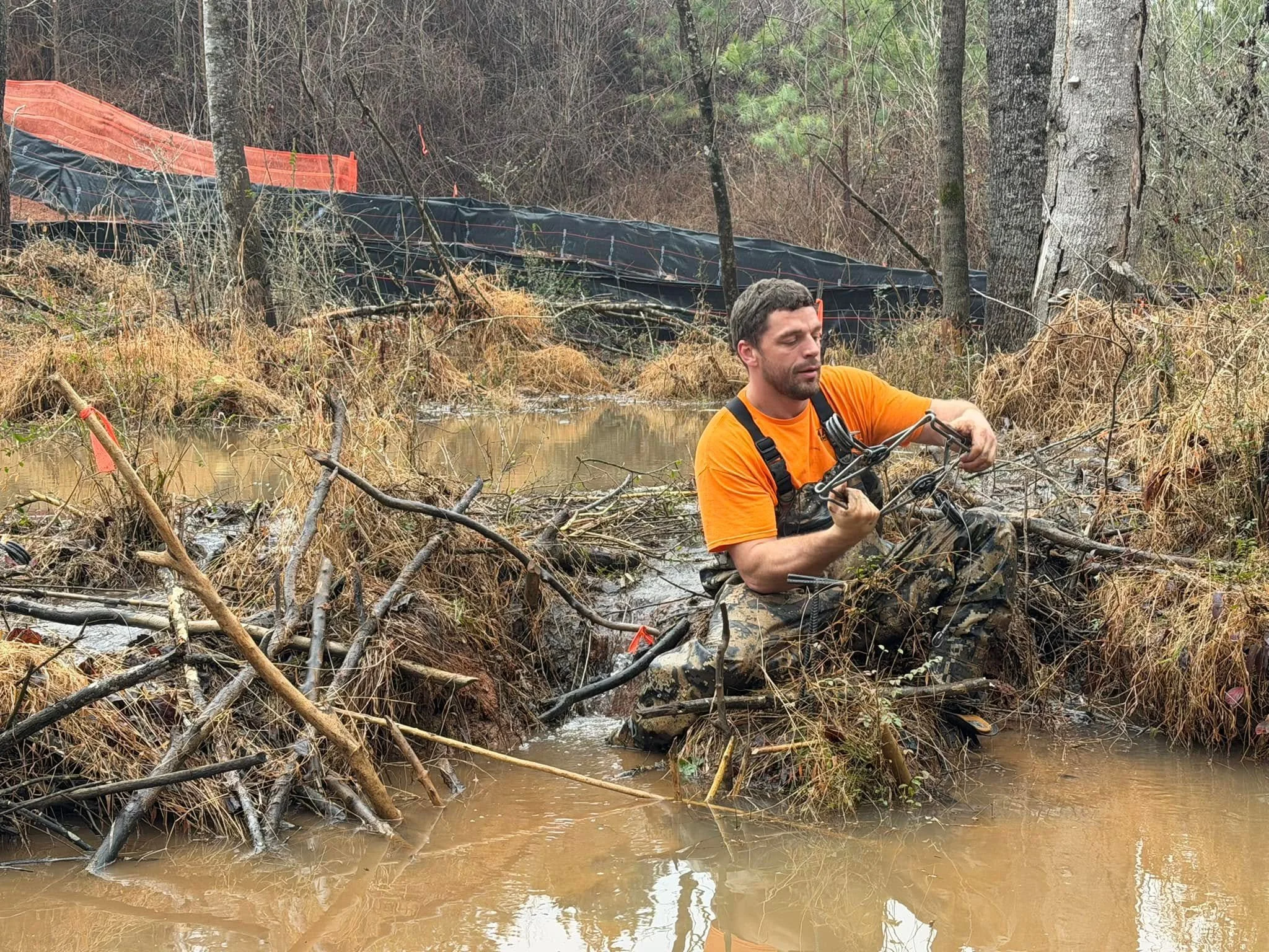 A man kneeling in a muddy creek, wearing an orange shirt and suspenders, surrounded by fallen branches and trees after a flood.