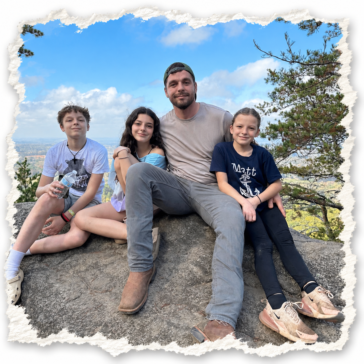 A man and three children sitting on a rock outdoors with trees and a scenic view of the sky and landscape in the background.