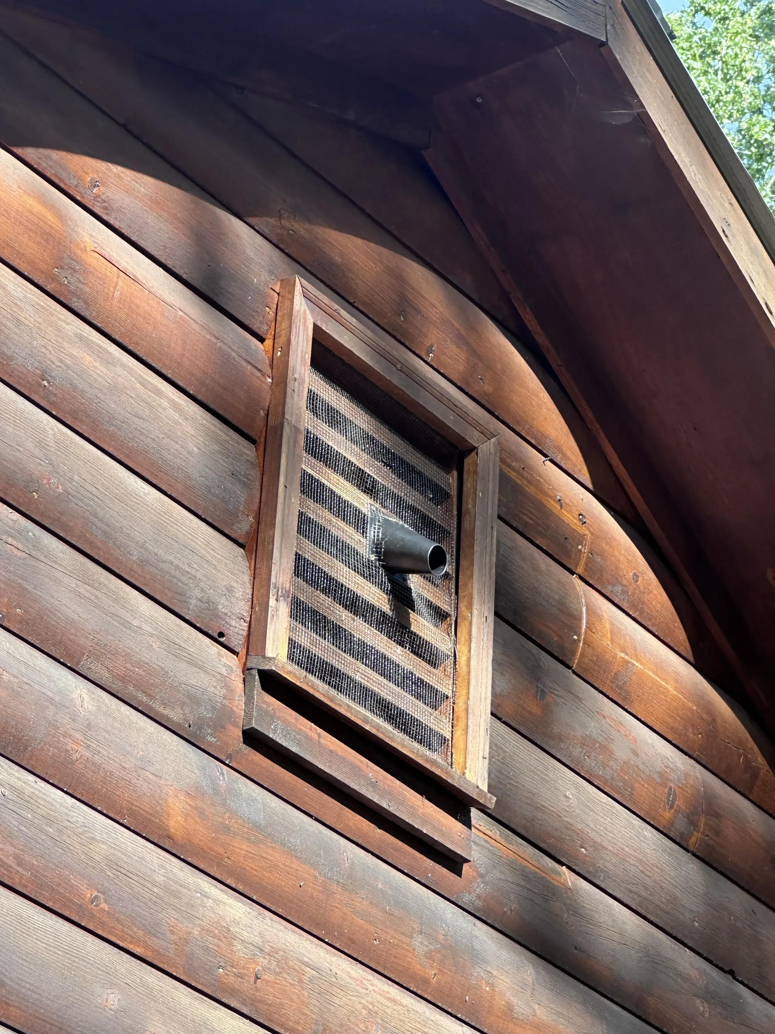 Close-up of a wooden building exterior with a small screened vent and a black pipe protruding from the wall, sunlight illuminating the wood.