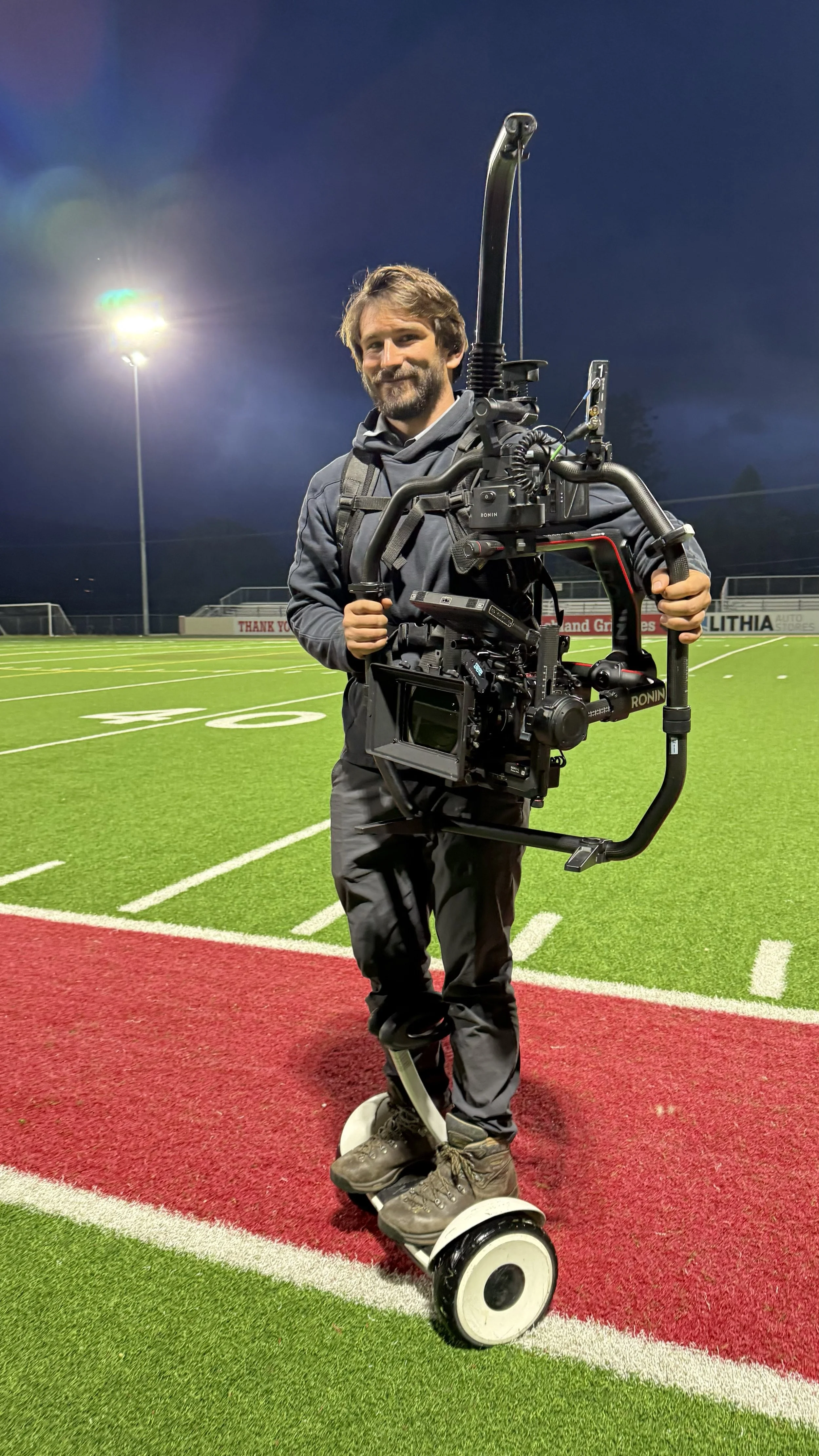 A man standing on a hoverboard on a football field holding a large professional camera stabilizer rig, with stadium lights and dark evening sky in the background.