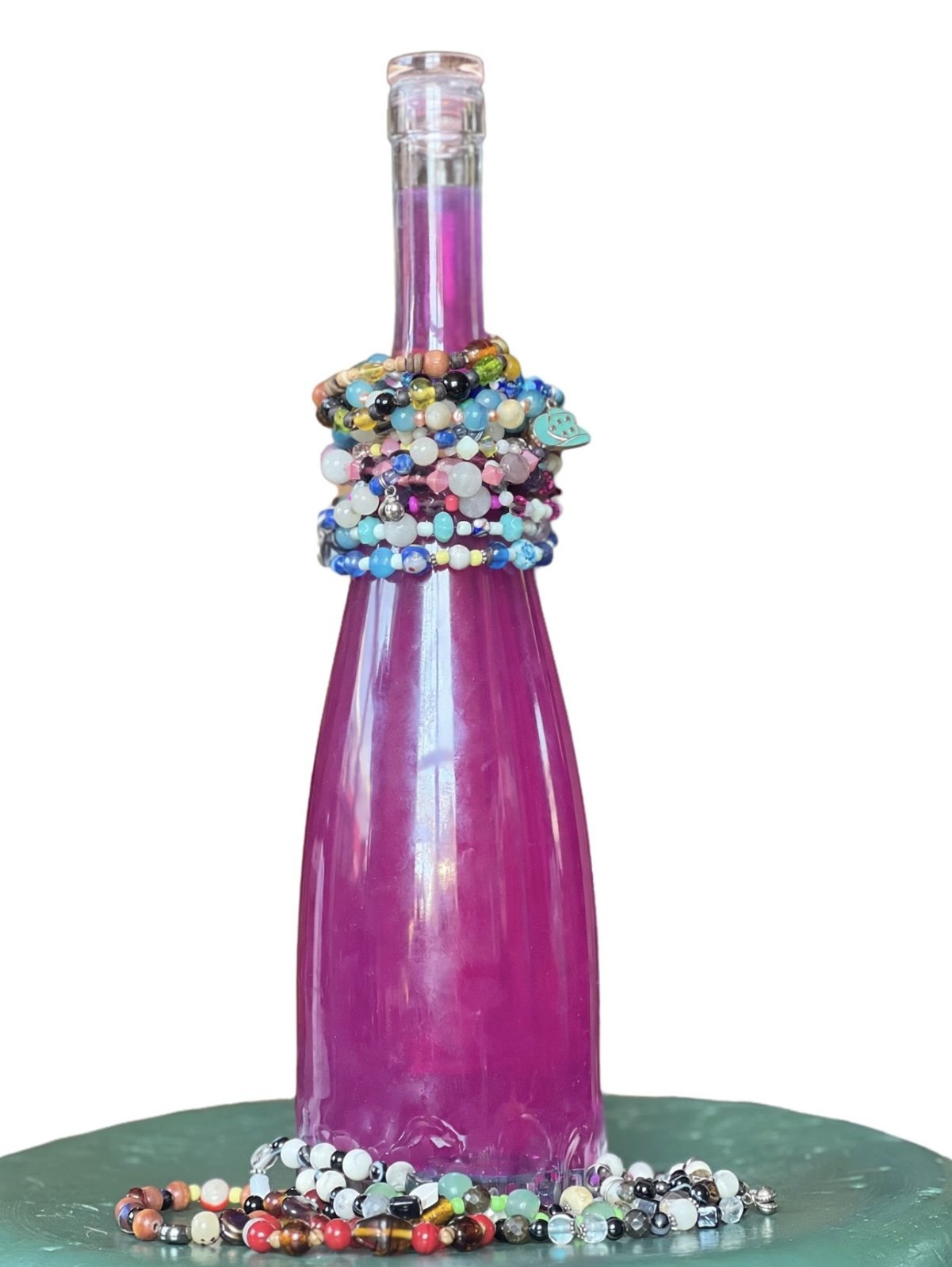 Pink glass bottle decorated with colorful beads, rings, and charms, placed on a green surface.