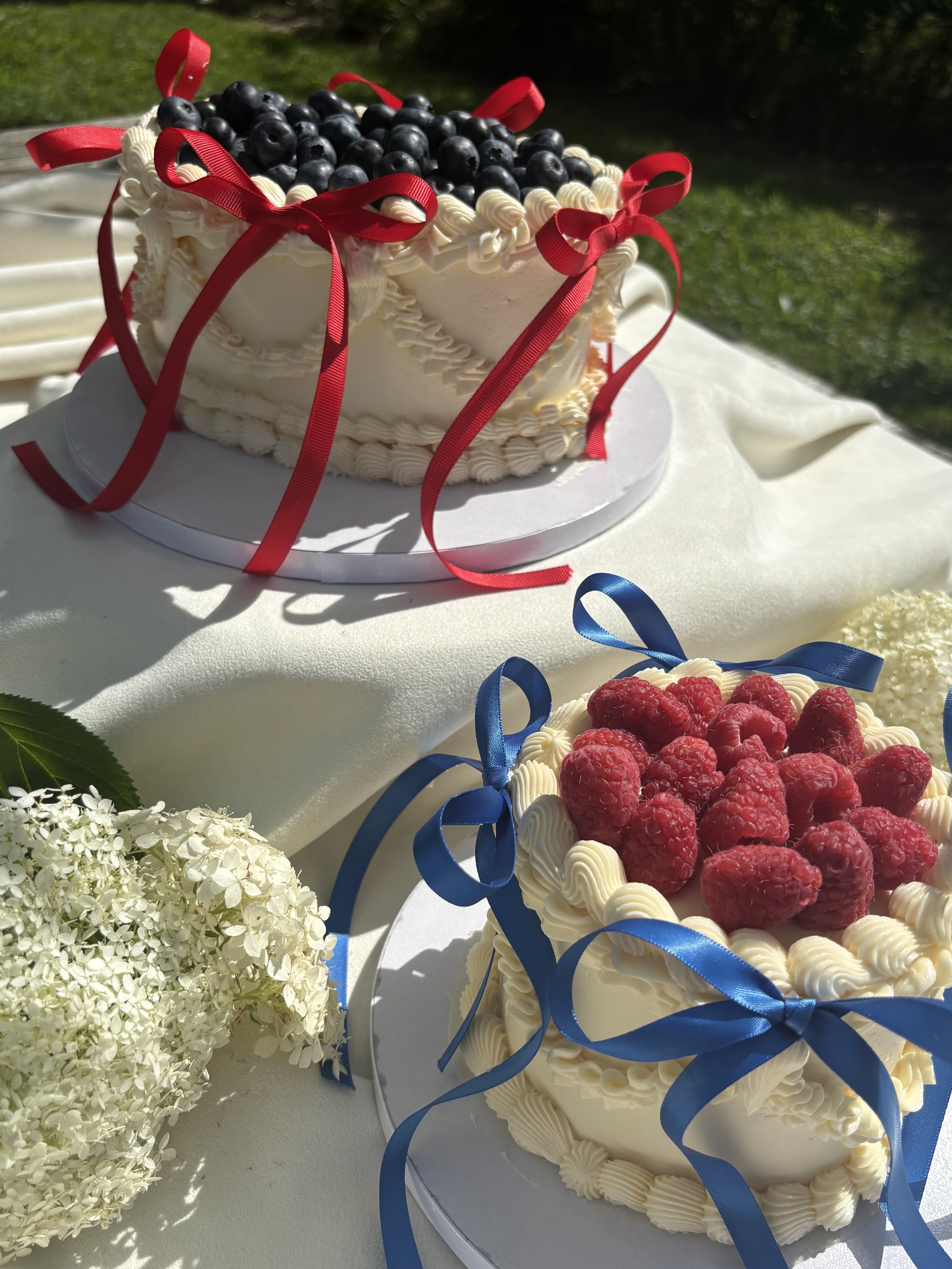 A white layered cake topped with blueberries and decorated with red ribbons, placed on a white table, with another similar cake decorated with raspberries and blue ribbons nearby, and white flower arrangements surrounding the setup.