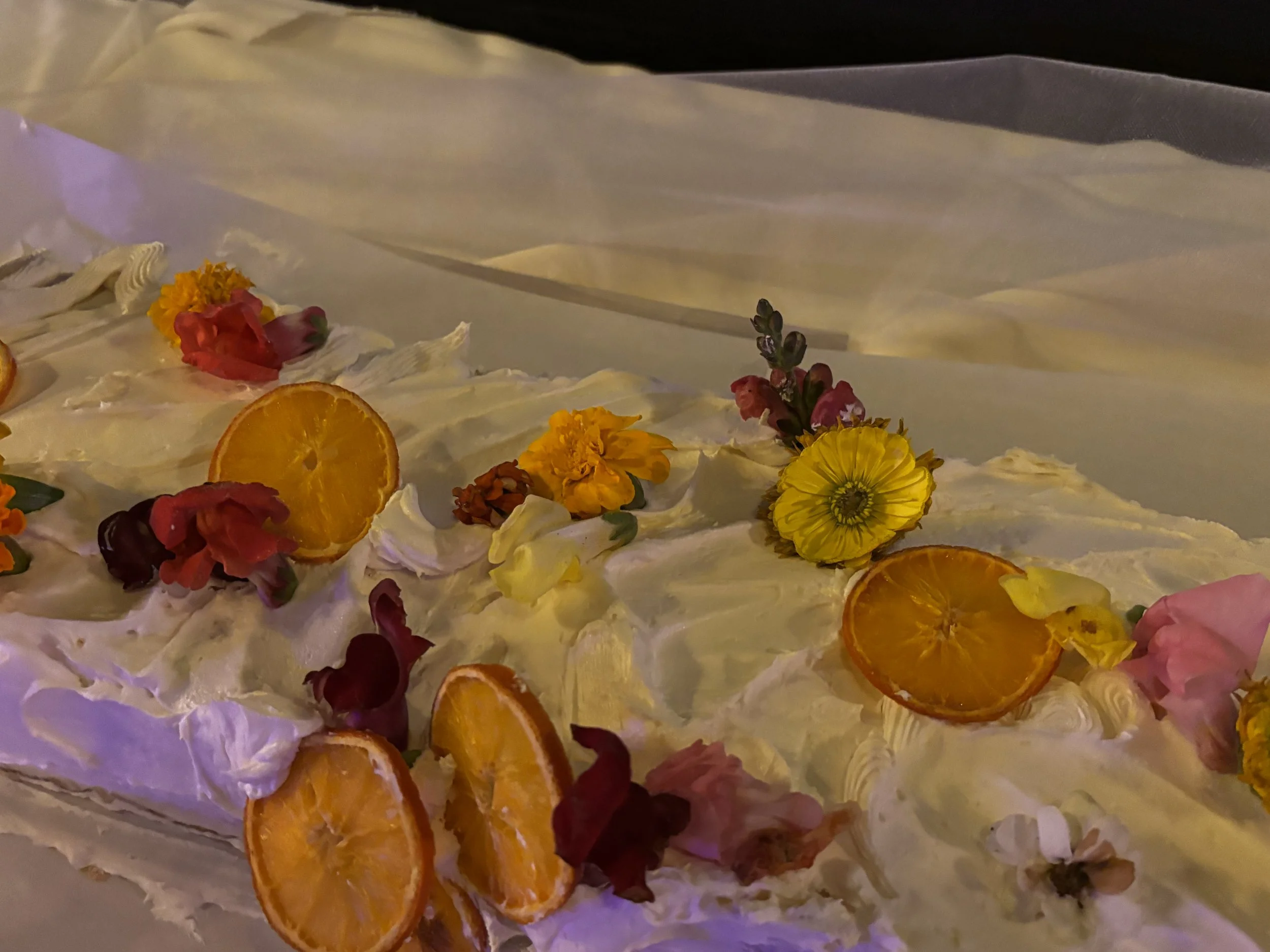 Close-up of a cake decorated with white frosting, orange slices, yellow and pink flowers, and purple flower petals.