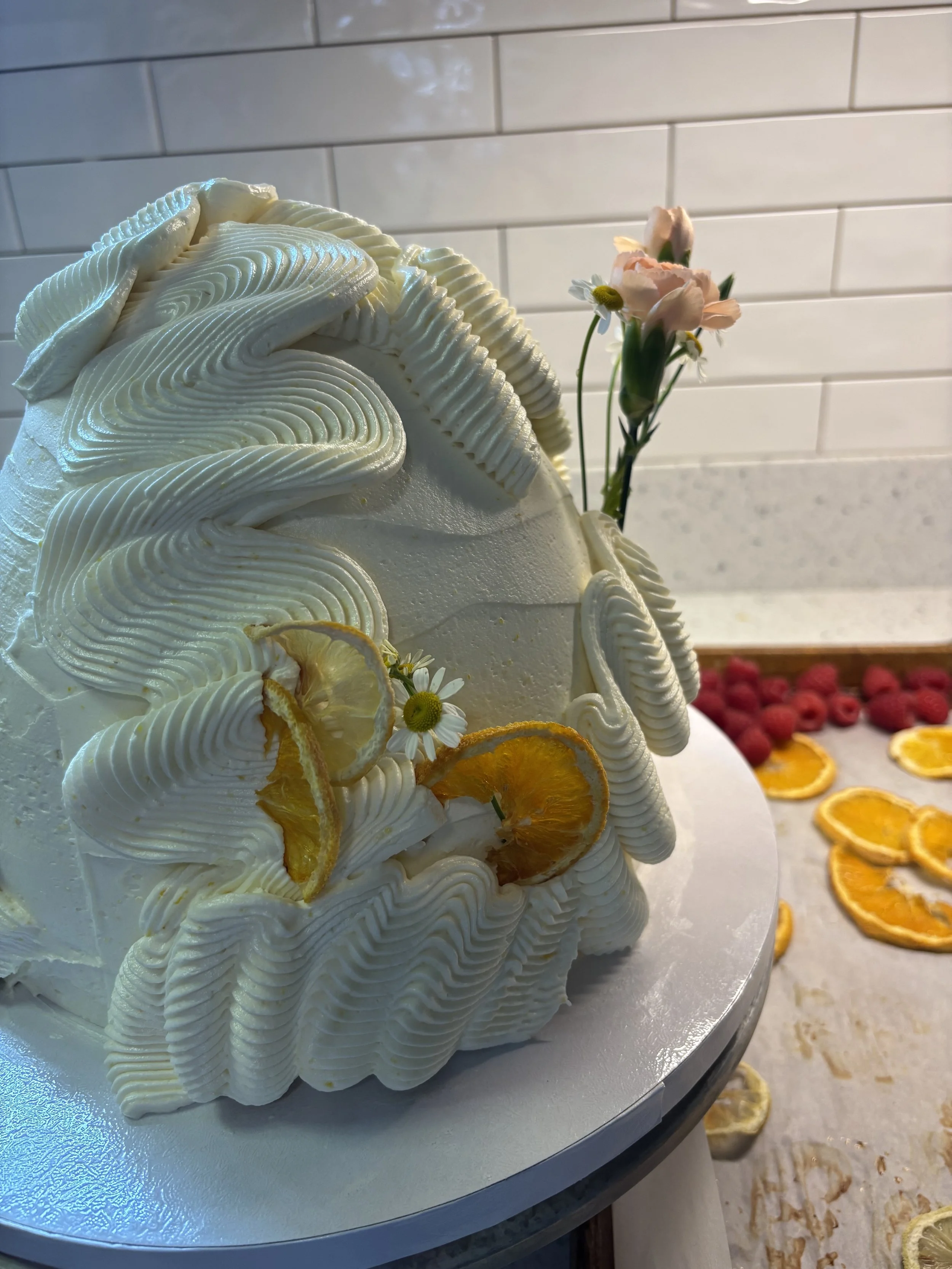A layered white cake decorated with Rolled piped icing, dried lemon slices, and small flowers with a pink flower and chamomile. The cake is on a silver cake board with fruit slices and berries scattered around on the surface and background.