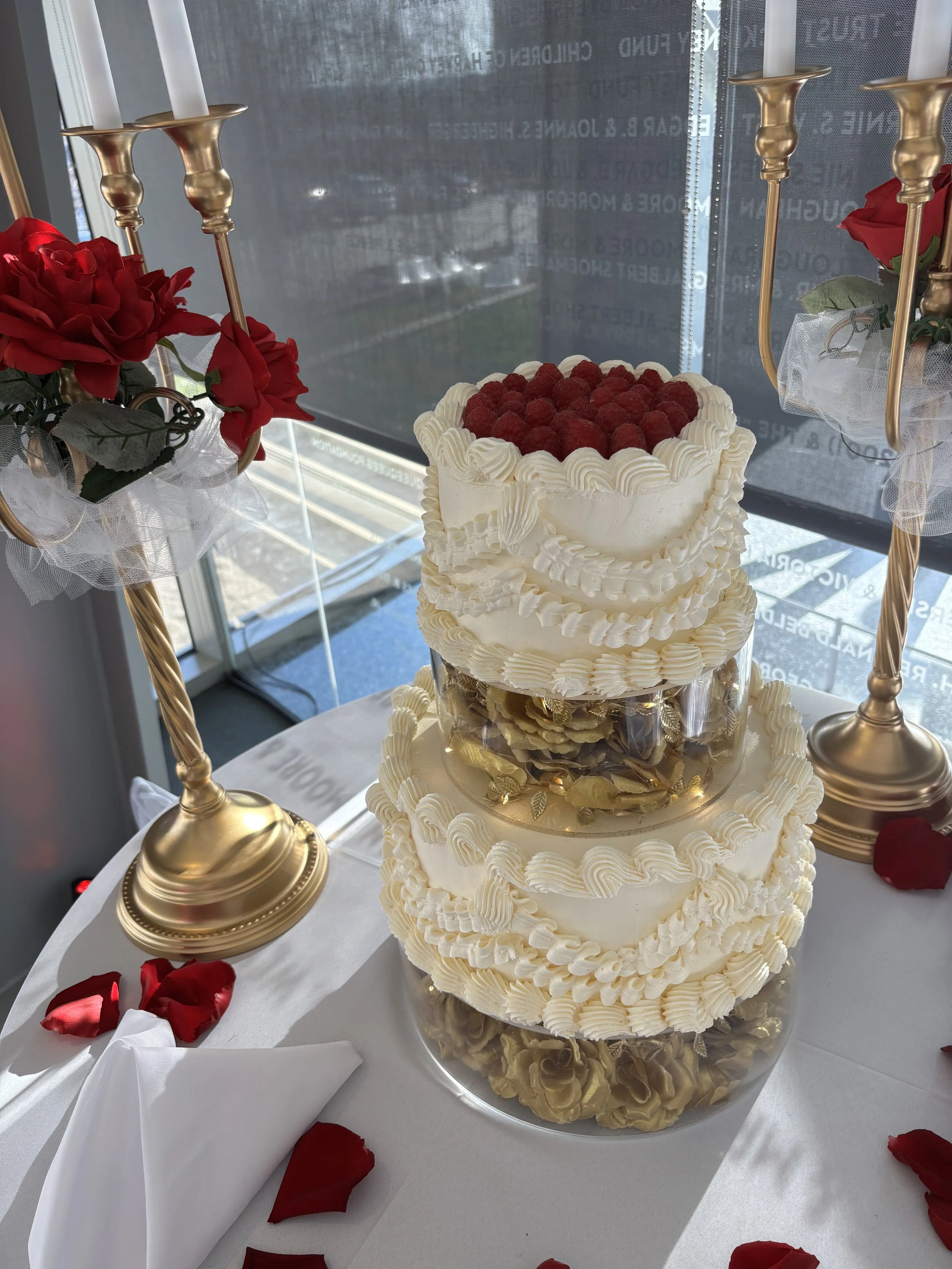 Three-tier wedding cake topped with raspberries, decorated with cream and flowers, surrounded by gold candleholders and scattered red rose petals on a white tablecloth.